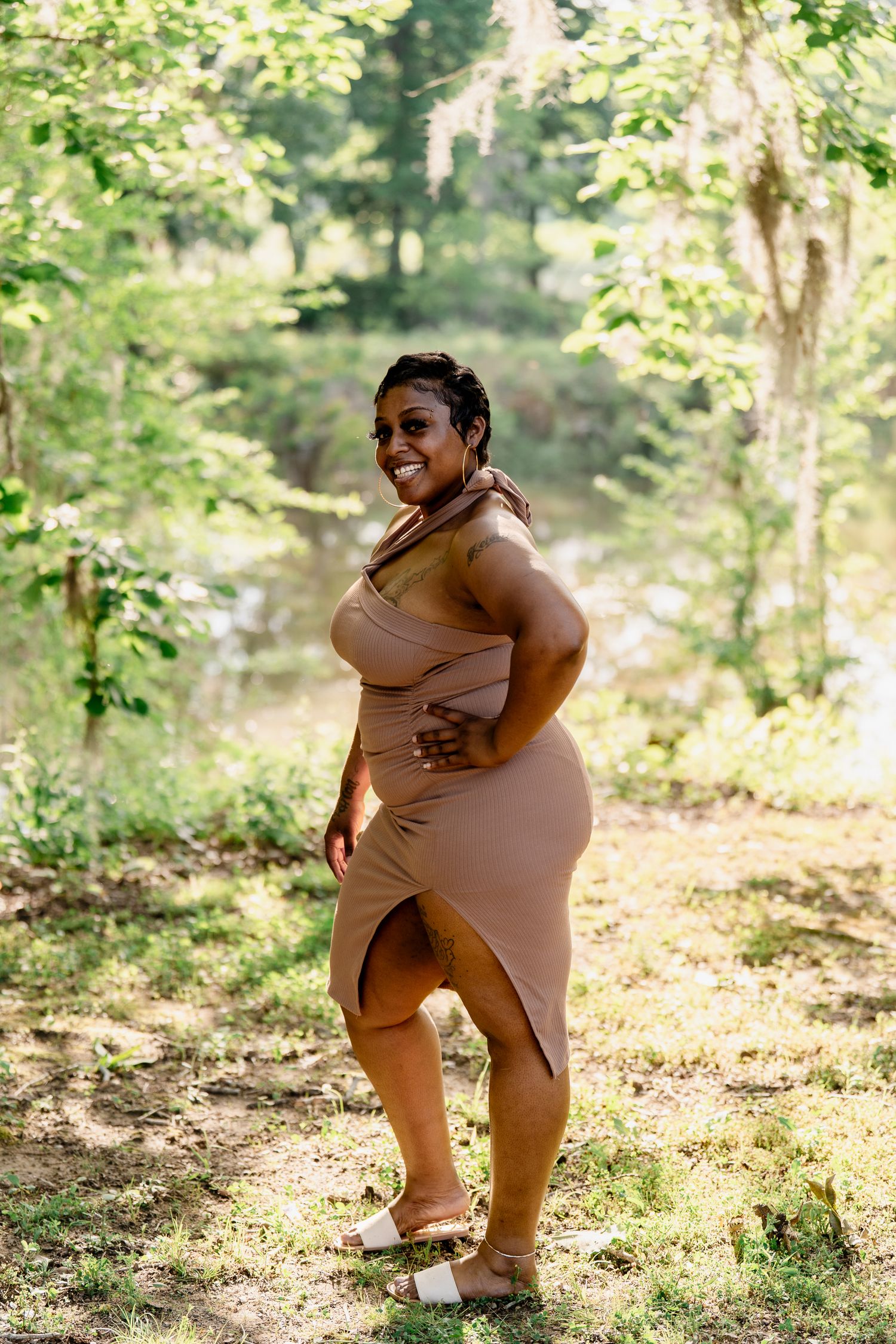 Woman in beige dress strikes a confident pose in a sun-dappled forest setting.