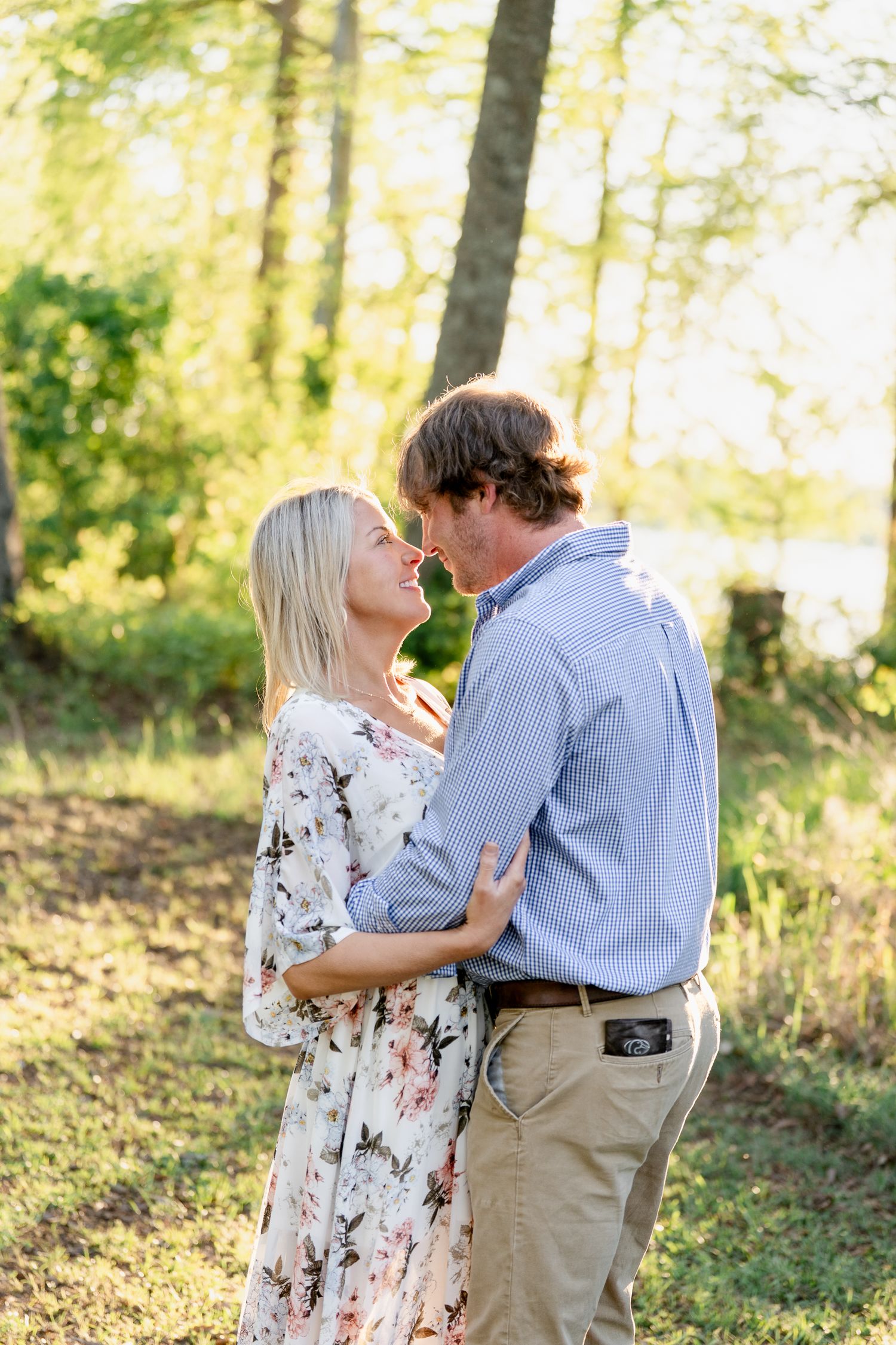 A romantic couple embraces in a sunlit forest setting, with her wearing a floral dress and him in blue shirt and khakis.