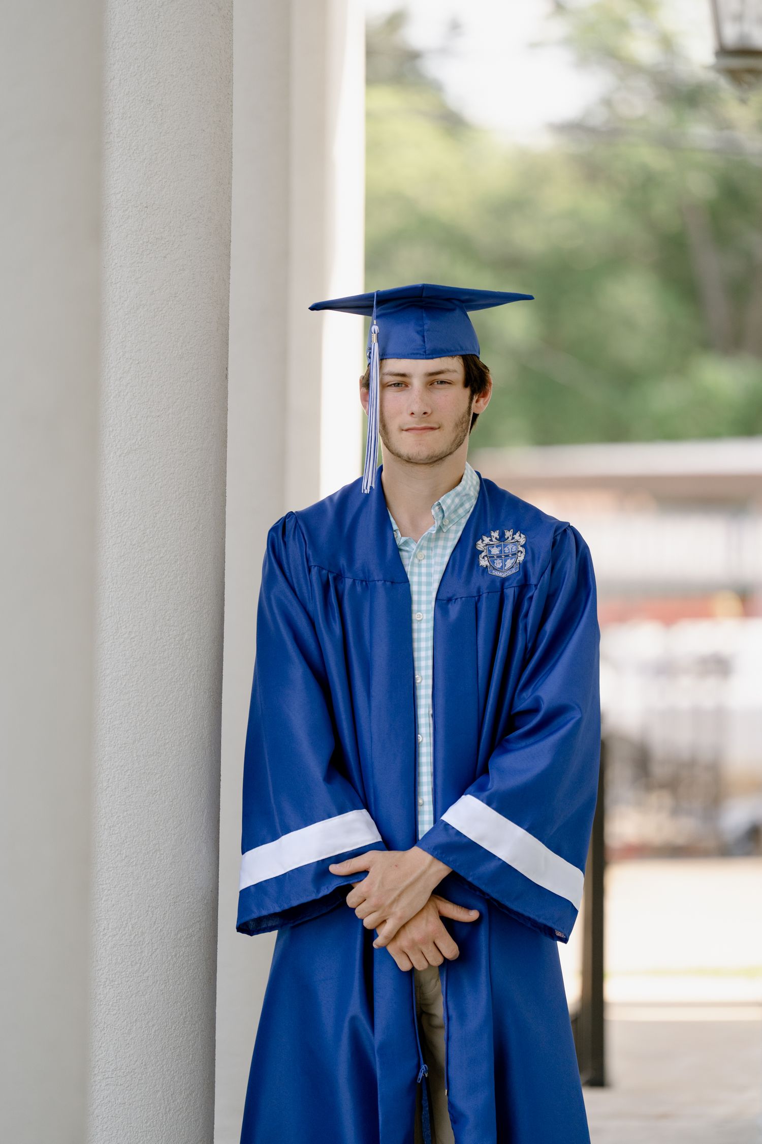 Graduate stands formally in blue graduation gown against white column background.