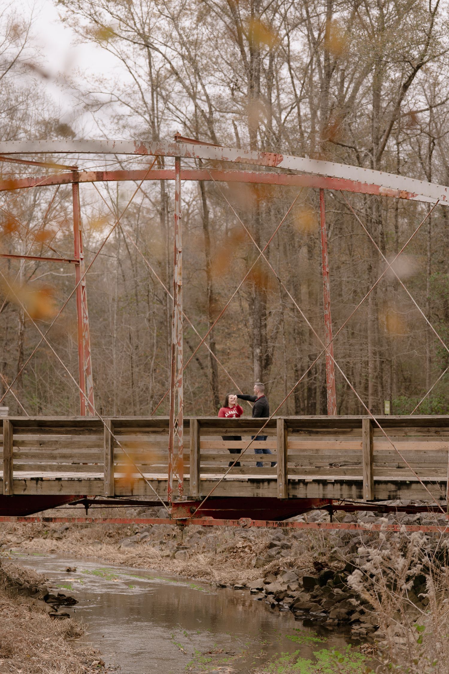 A rustic red metal bridge spans over a creek with bare winter trees in the background.
