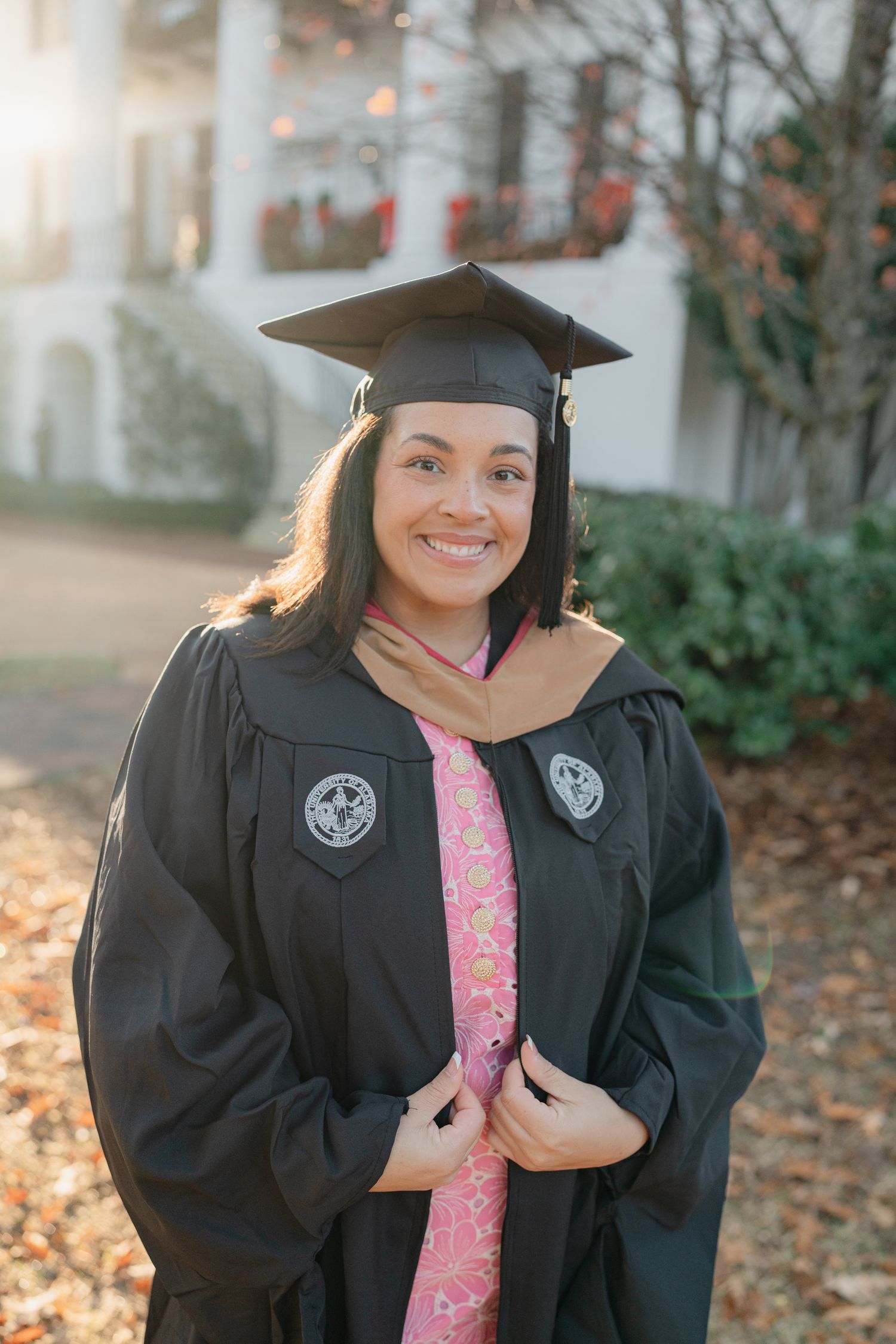 A graduate in black cap and gown poses outdoors with a pink sash against an autumn backdrop.