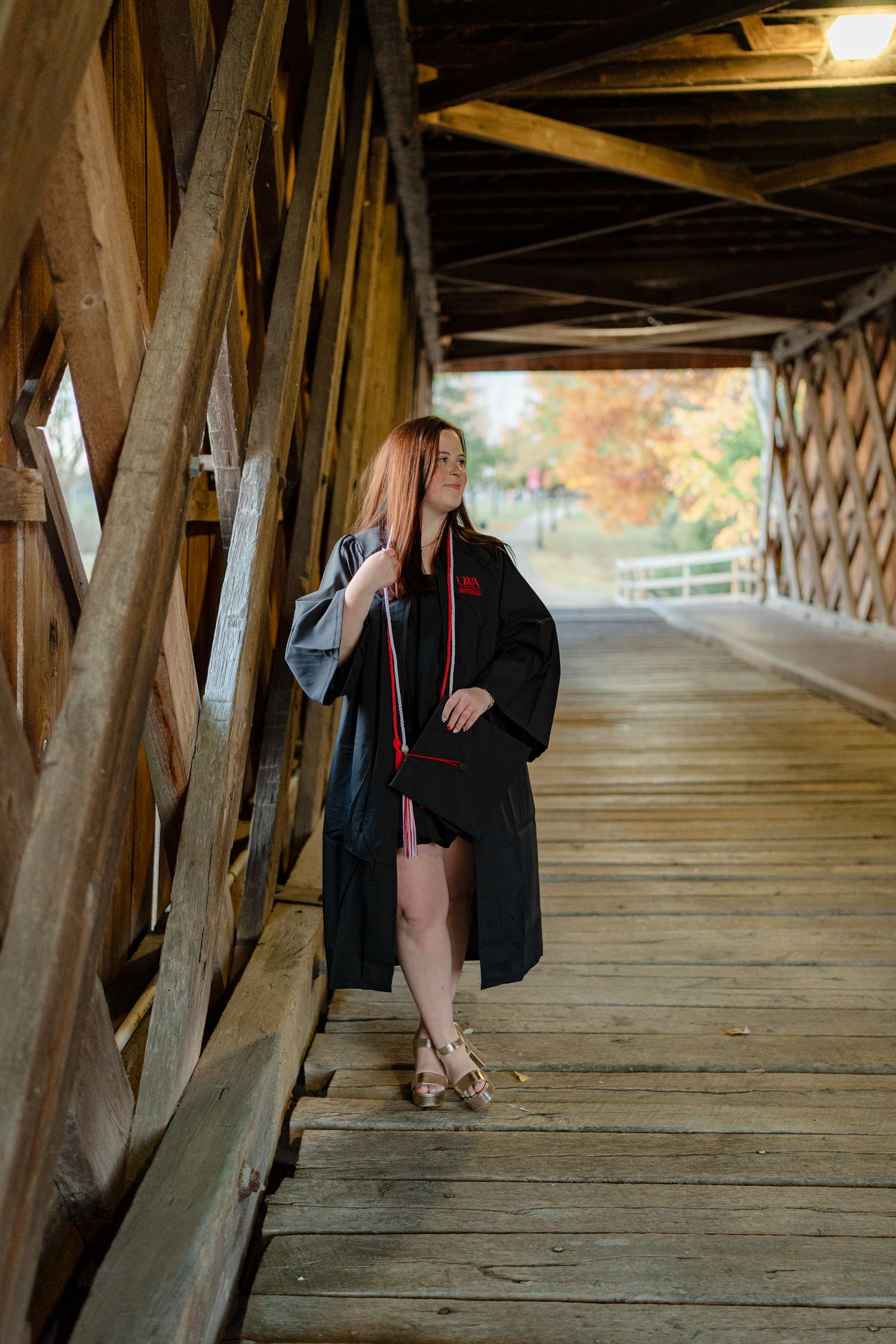 Graduate in black academic regalia poses inside a rustic wooden covered bridge during golden hour.