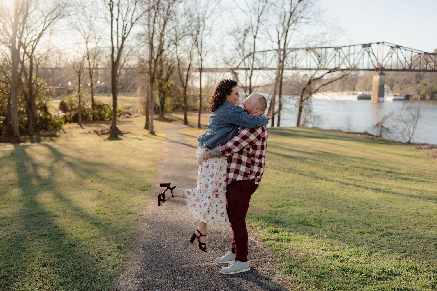 A romantic couple embraces near a rustic metal bridge at sunset in a serene park setting.