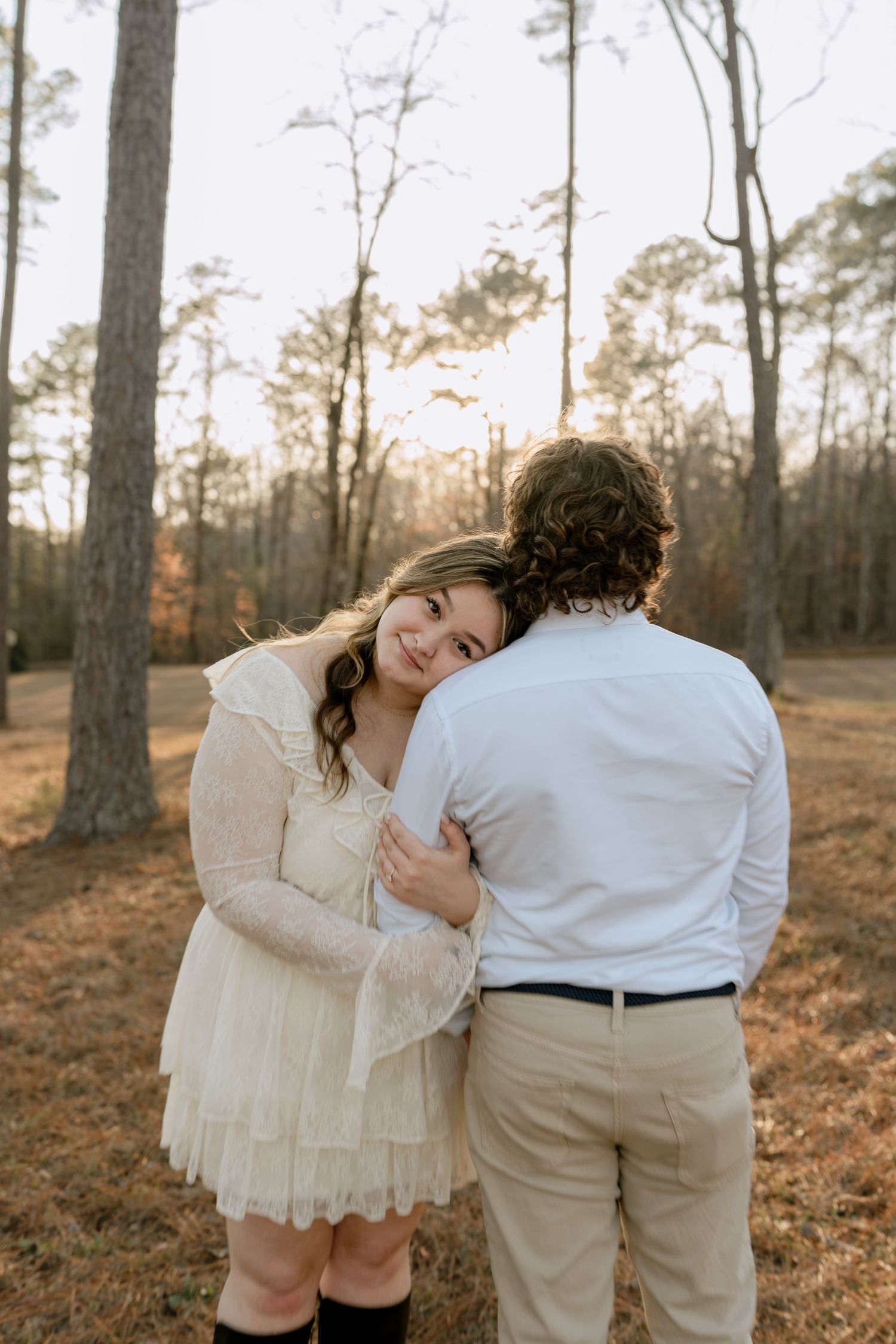 Smiling woman in lace mini dress hugging her partner among tall pine trees at sunset.