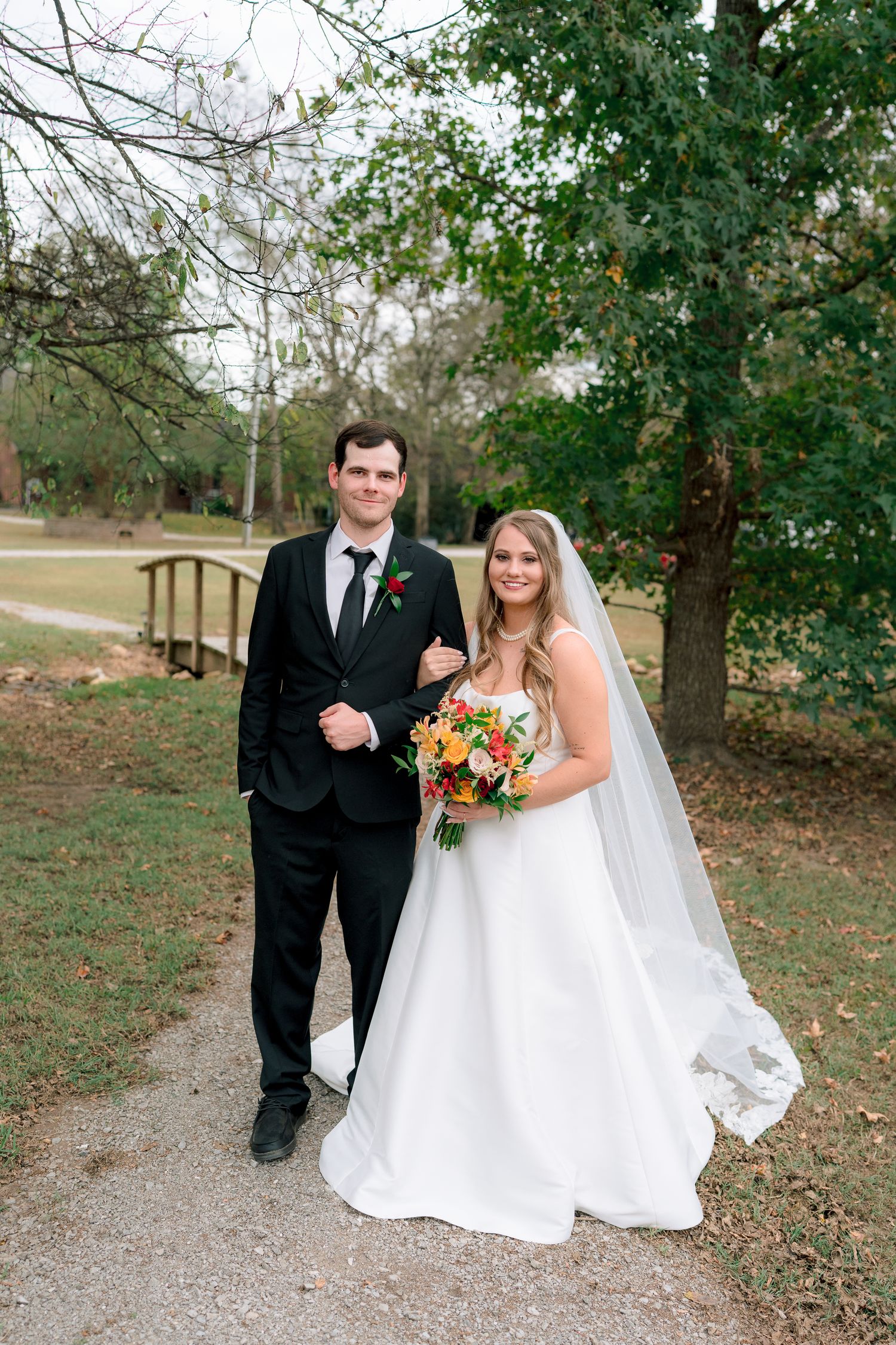 Bride in white gown with veil holding floral bouquet stands beside groom in black suit on garden path.
