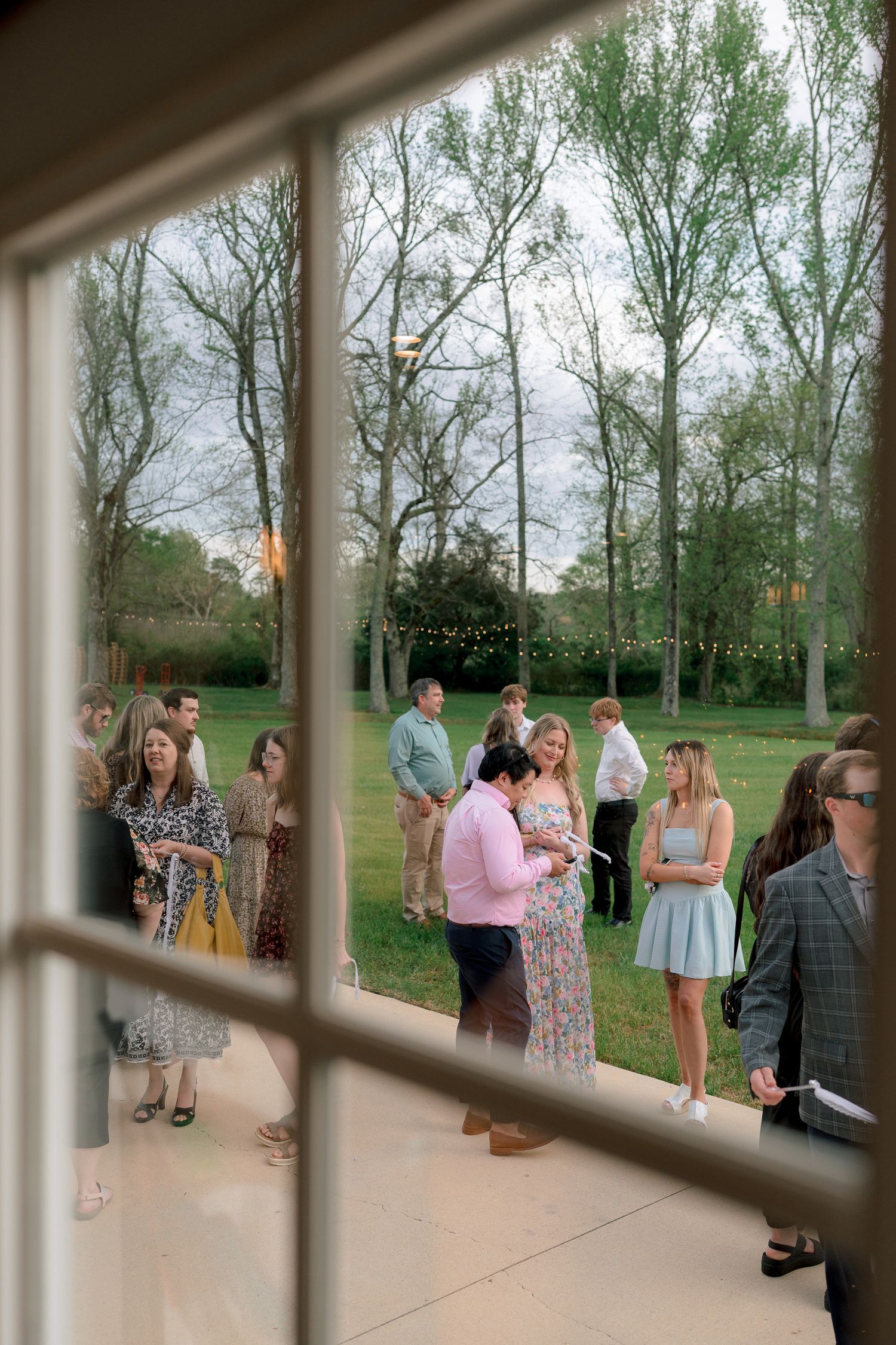 Wedding guests mingling on a patio viewed through a window, surrounded by trees with string lights at dusk.