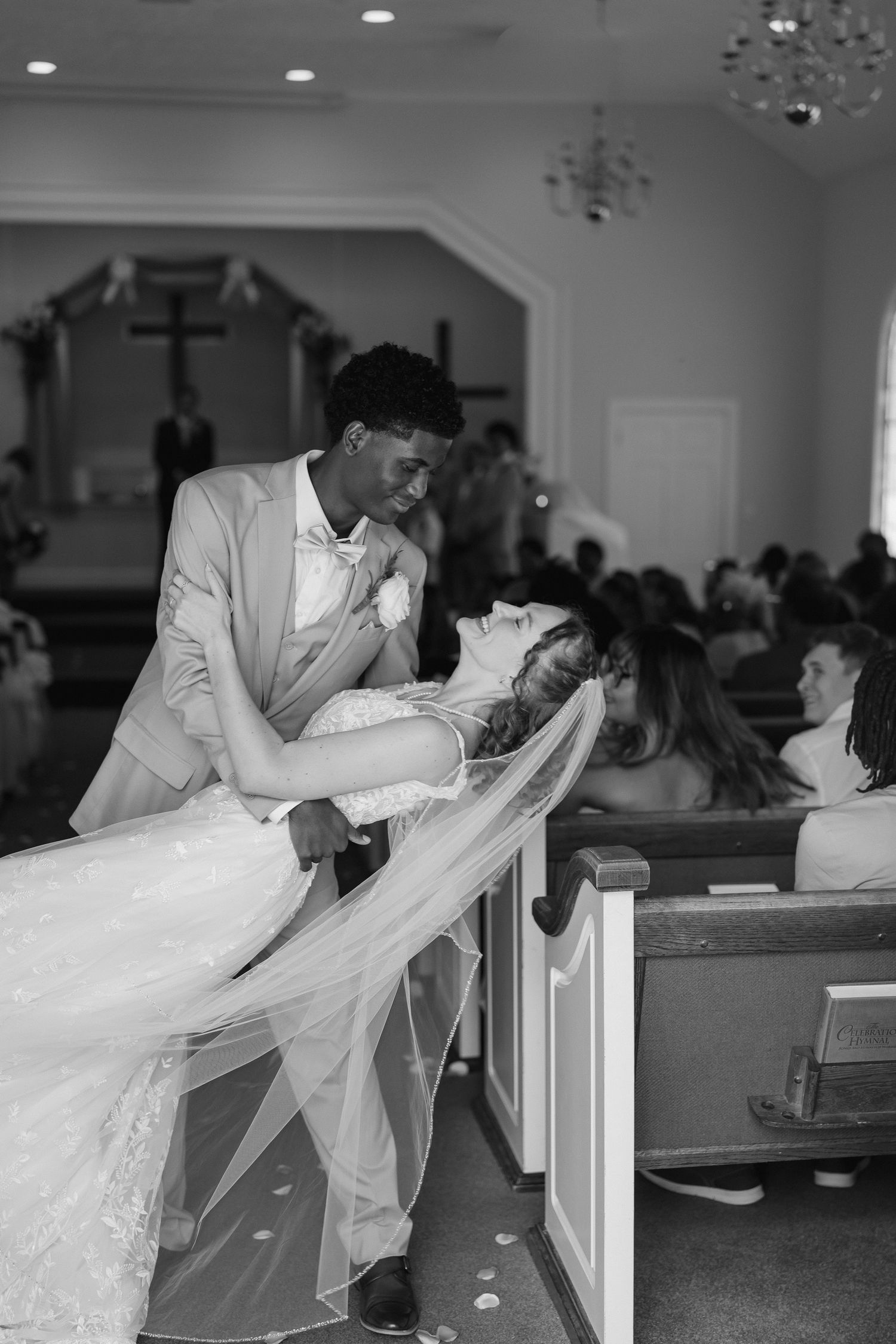 Groom dips bride in church aisle during wedding ceremony, black and white photo with guests seated in pews.