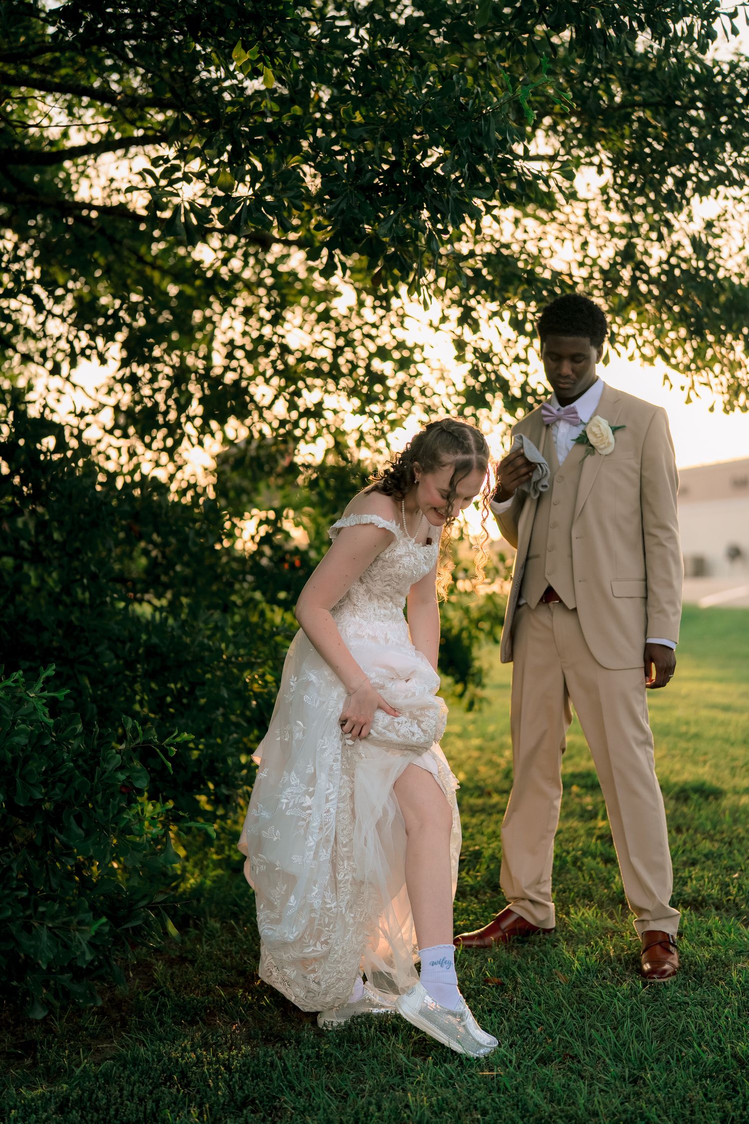 Bride lifting lace wedding dress to reveal white sneakers while groom watches under tree.