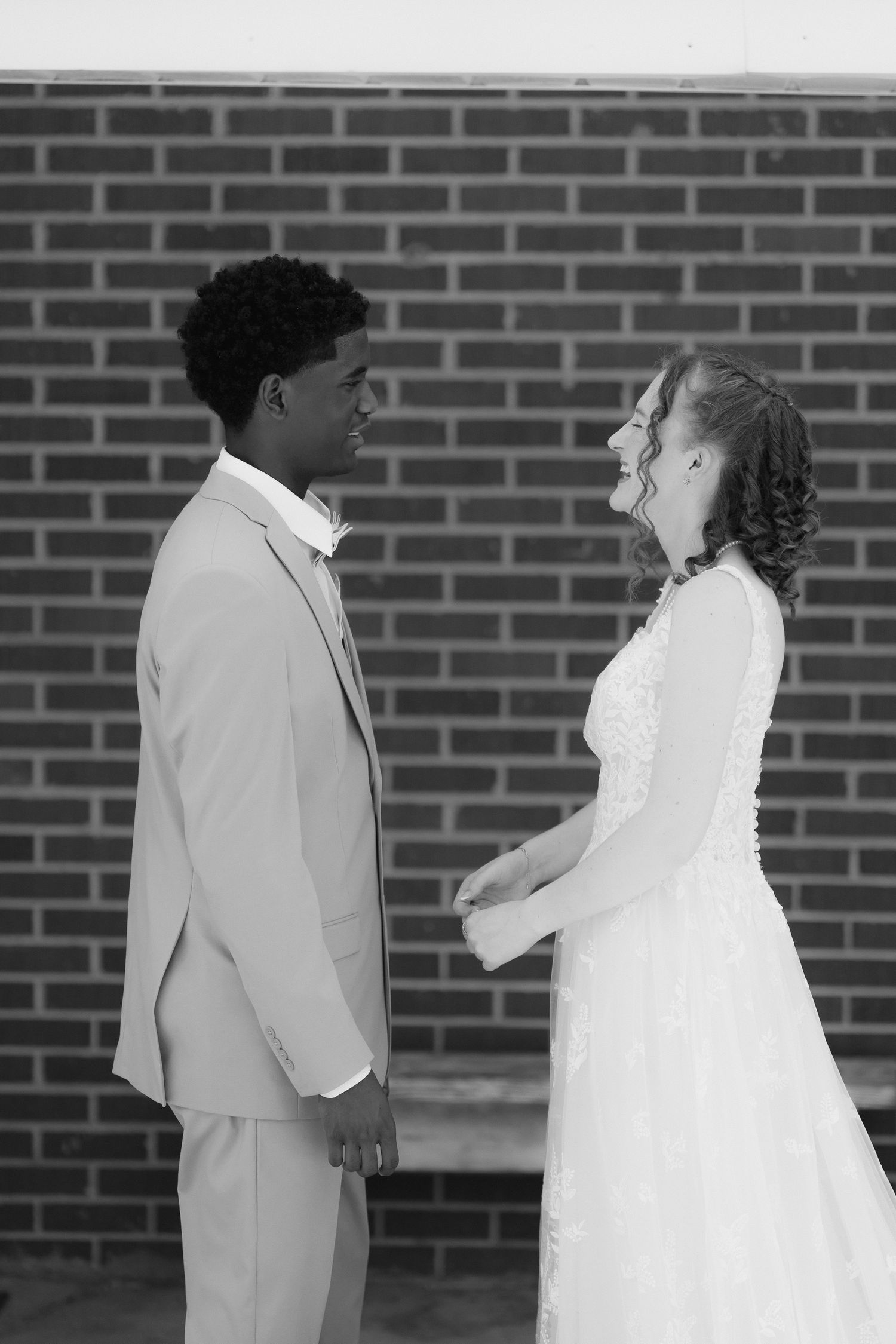 Black and white photo of smiling bride and groom holding hands outside brick wall on their wedding day.
