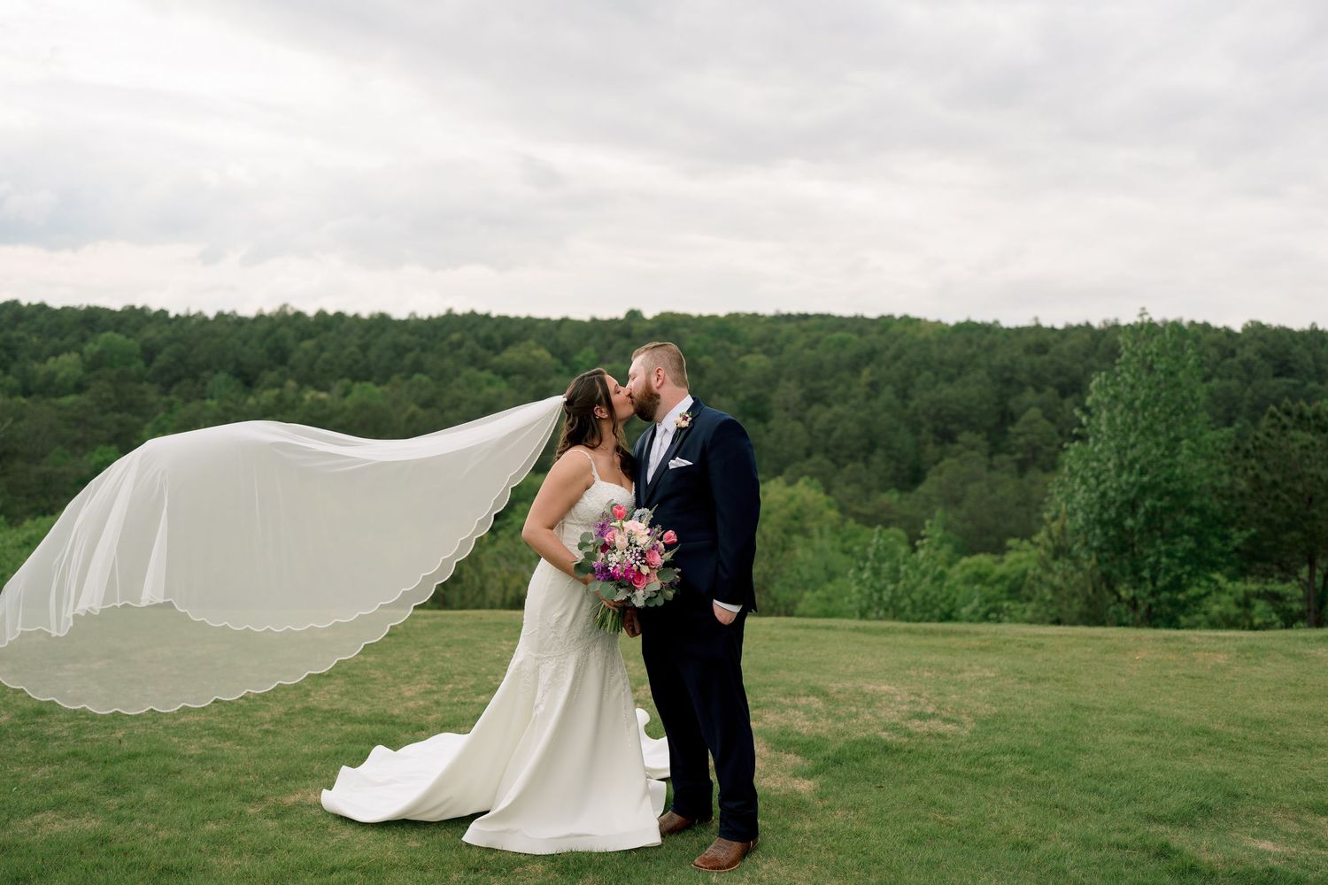 Bride and groom share a romantic kiss as her veil billows in the wind on a scenic green hillside.