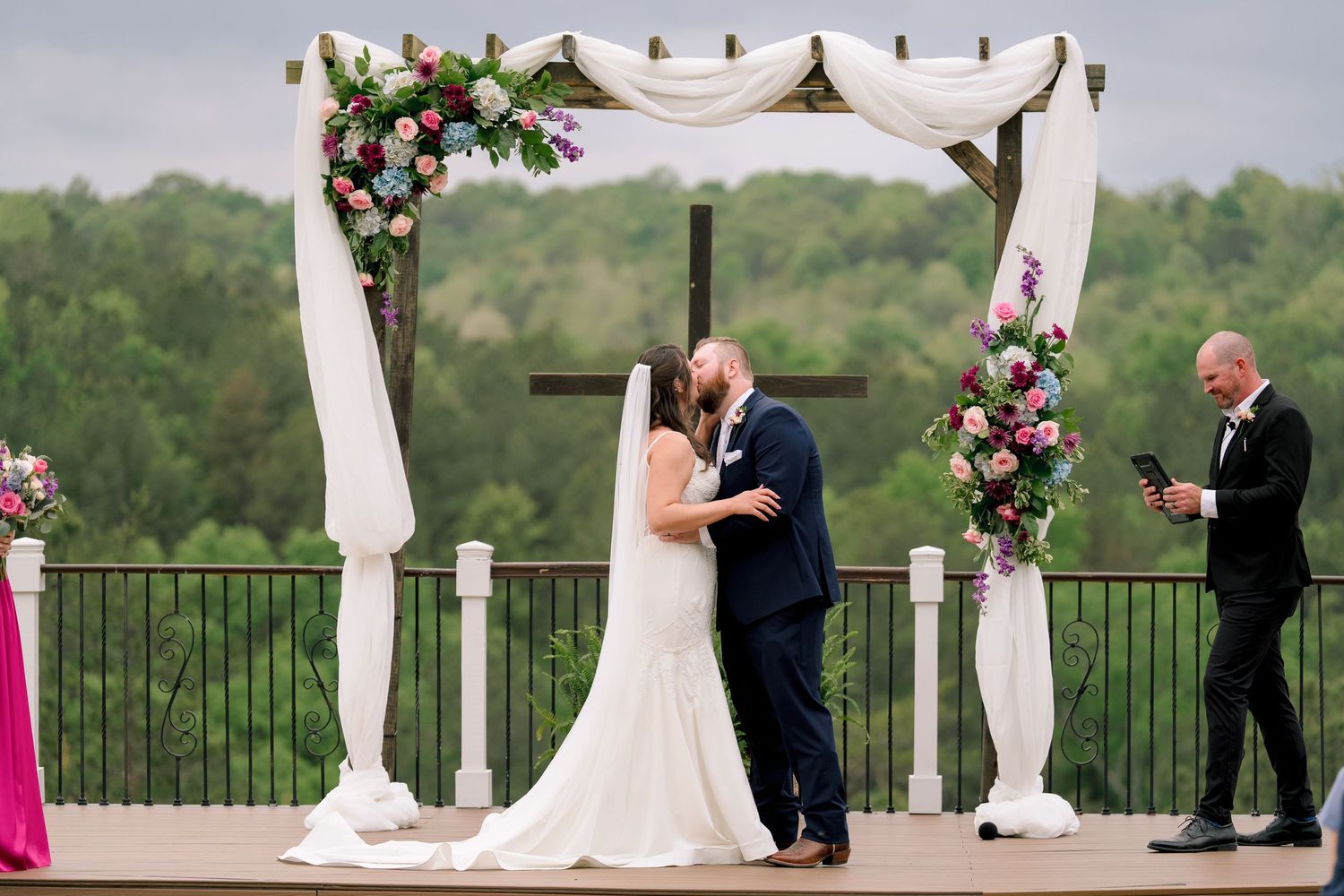 Bride and groom share first kiss under floral wedding arch with white draping and cross backdrop outdoors.
