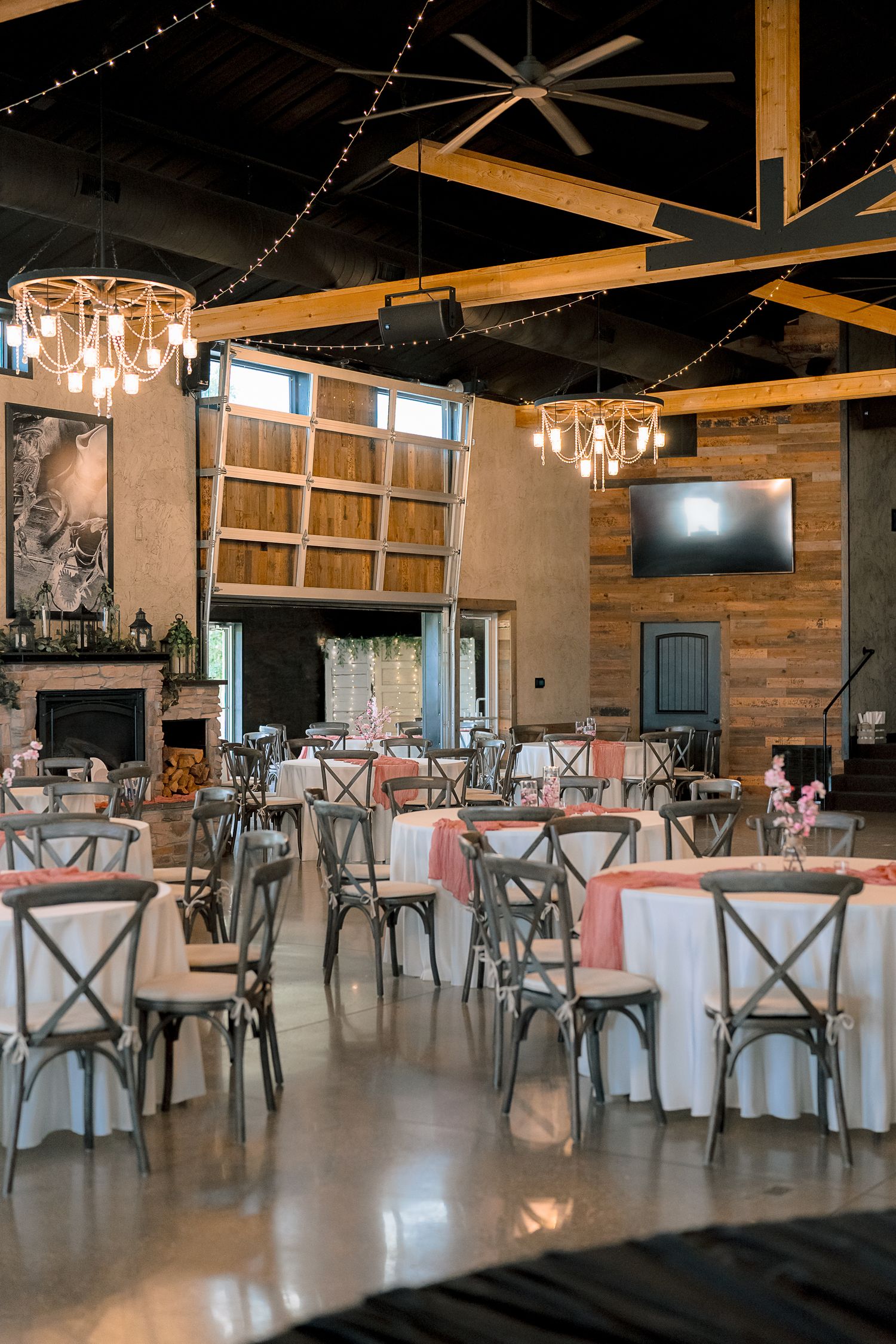 Elegant wedding reception venue with round tables, pink linens, cross-back chairs, chandeliers, and rustic wood beam ceiling.