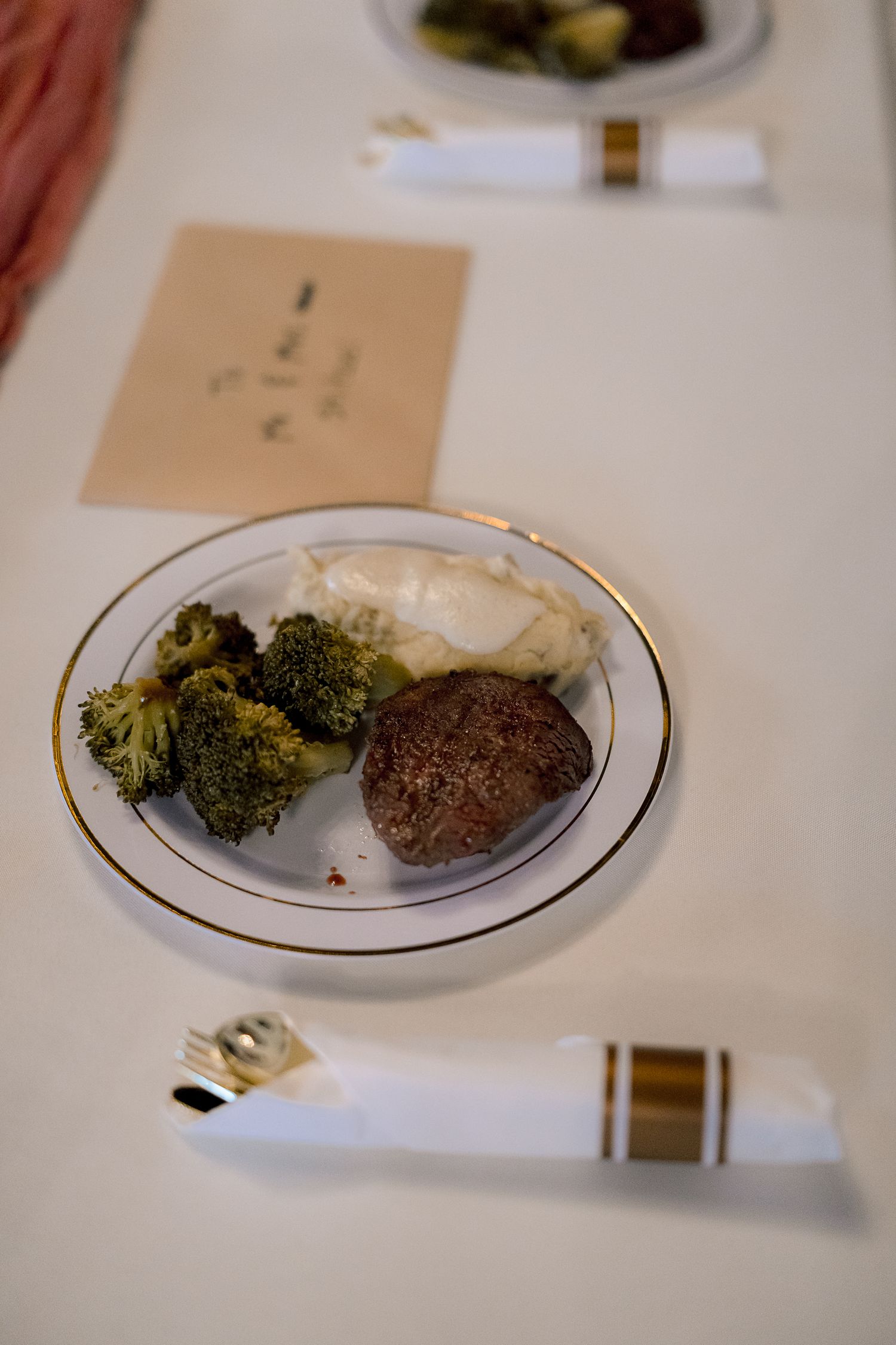 An elegant wedding reception dinner plate with filet mignon, broccoli, and mashed potatoes on a gold-rimmed dish.