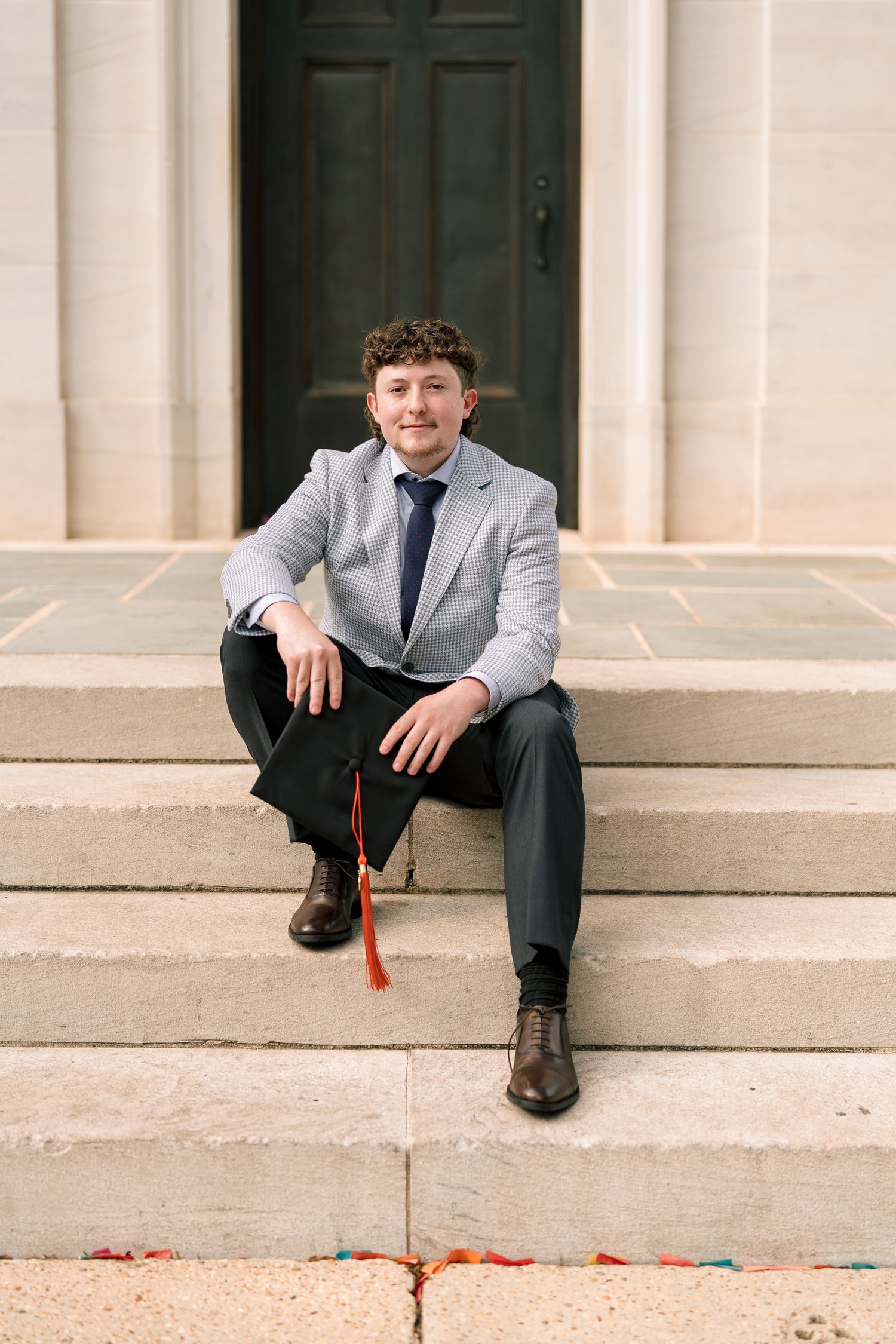 Young man in gray suit sitting on stone steps holding graduation cap with red tassel.