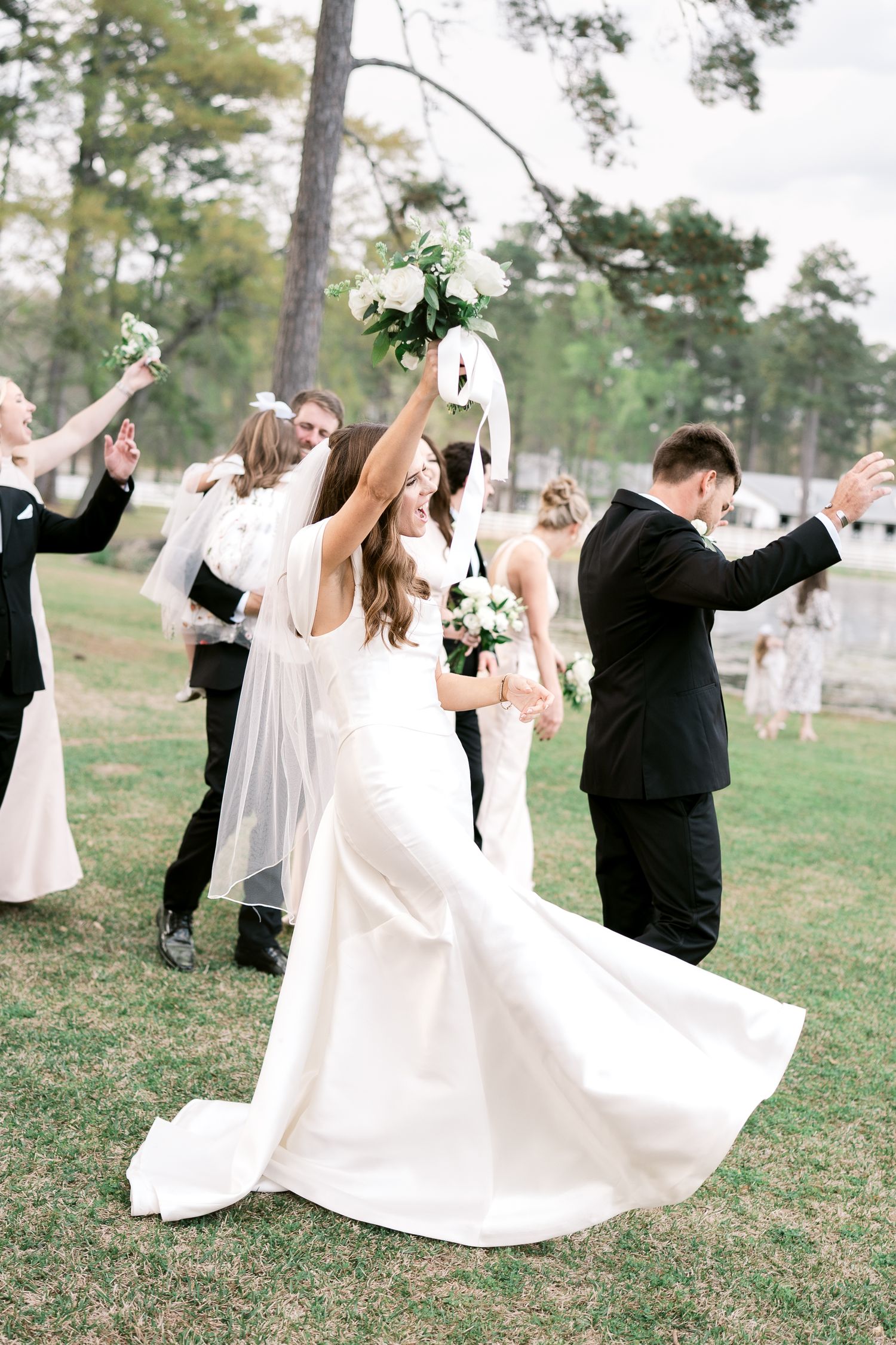 Wedding party celebrates joyfully on a grassy lawn surrounded by tall pine trees.
