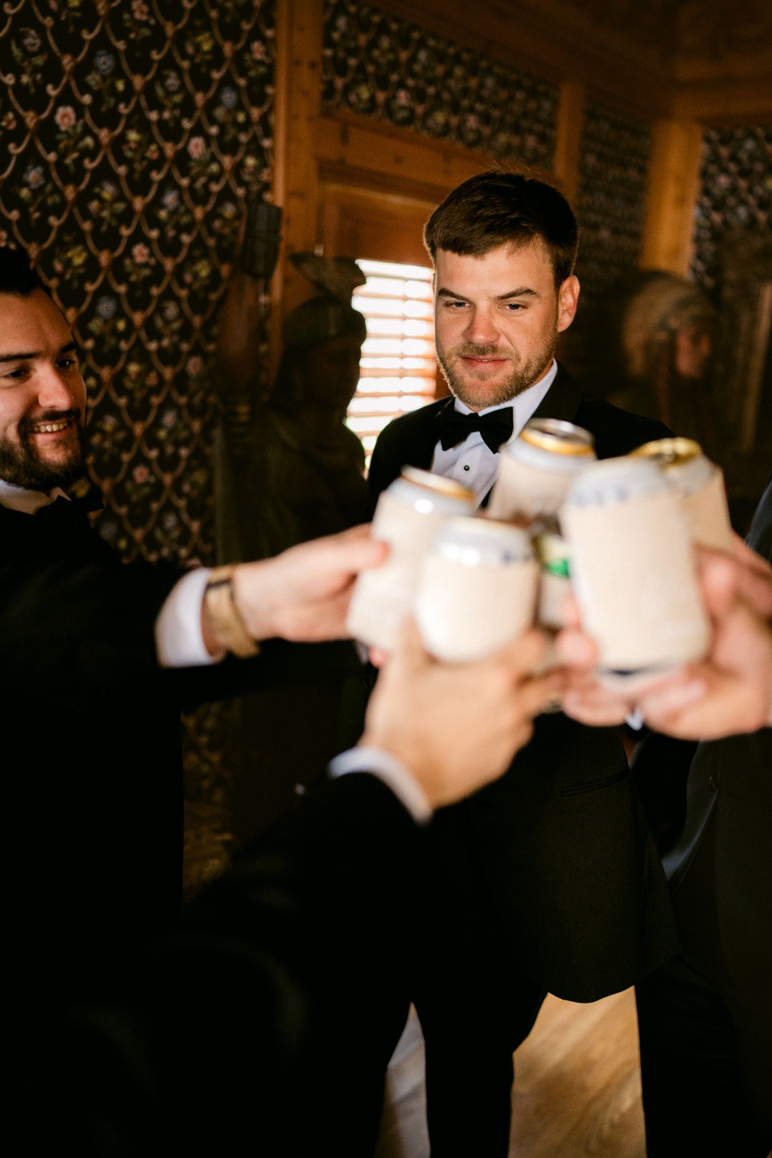 Groomsmen in tuxedos toast with white cups during a pre-wedding celebration.