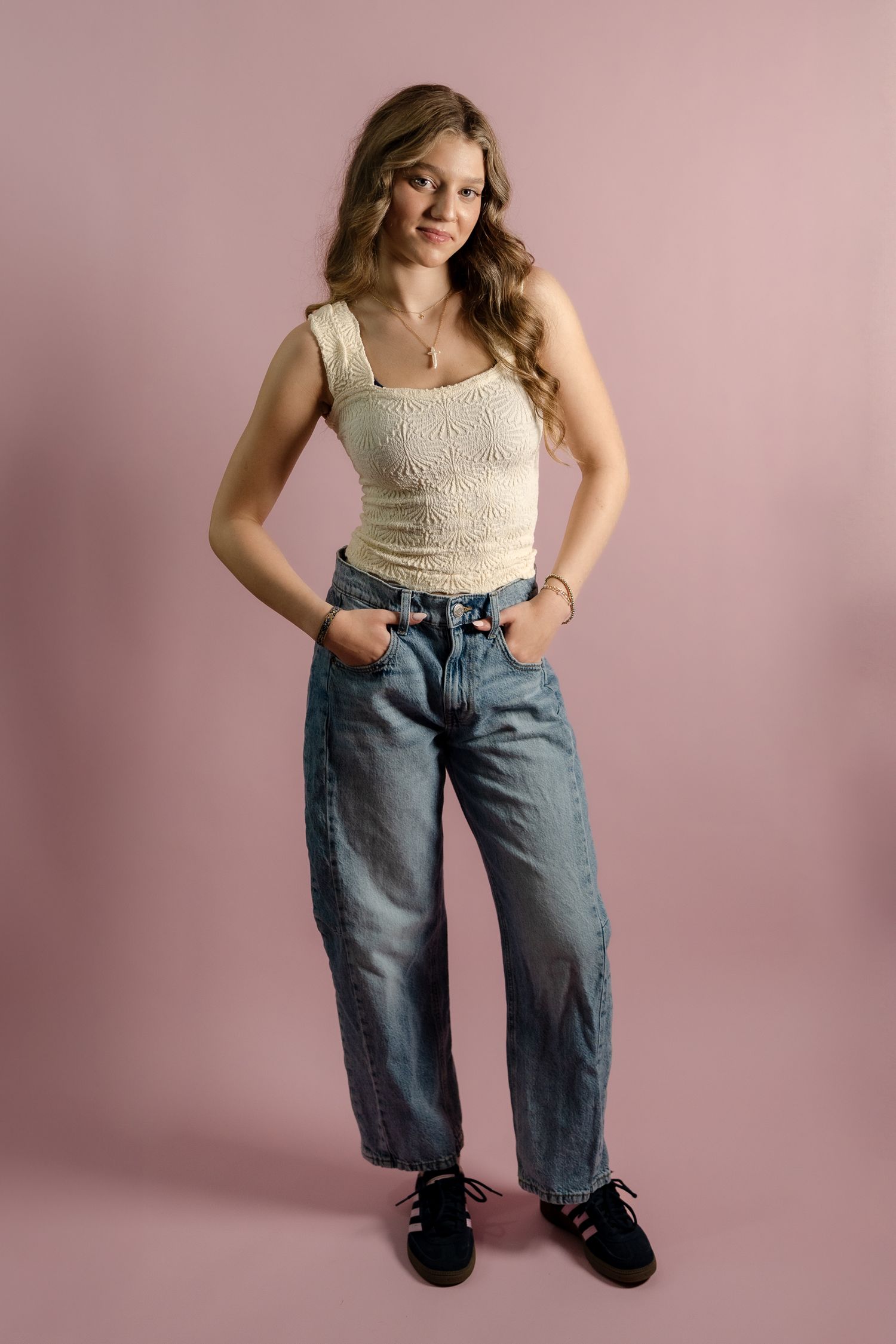 Fashion model poses in casual outfit with cream tank top and baggy jeans against pink backdrop.