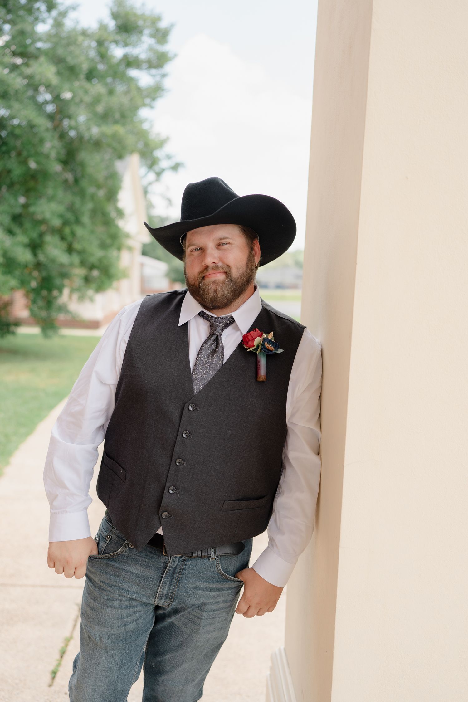 A person in western attire including a black cowboy hat and vest leans against a white pillar.