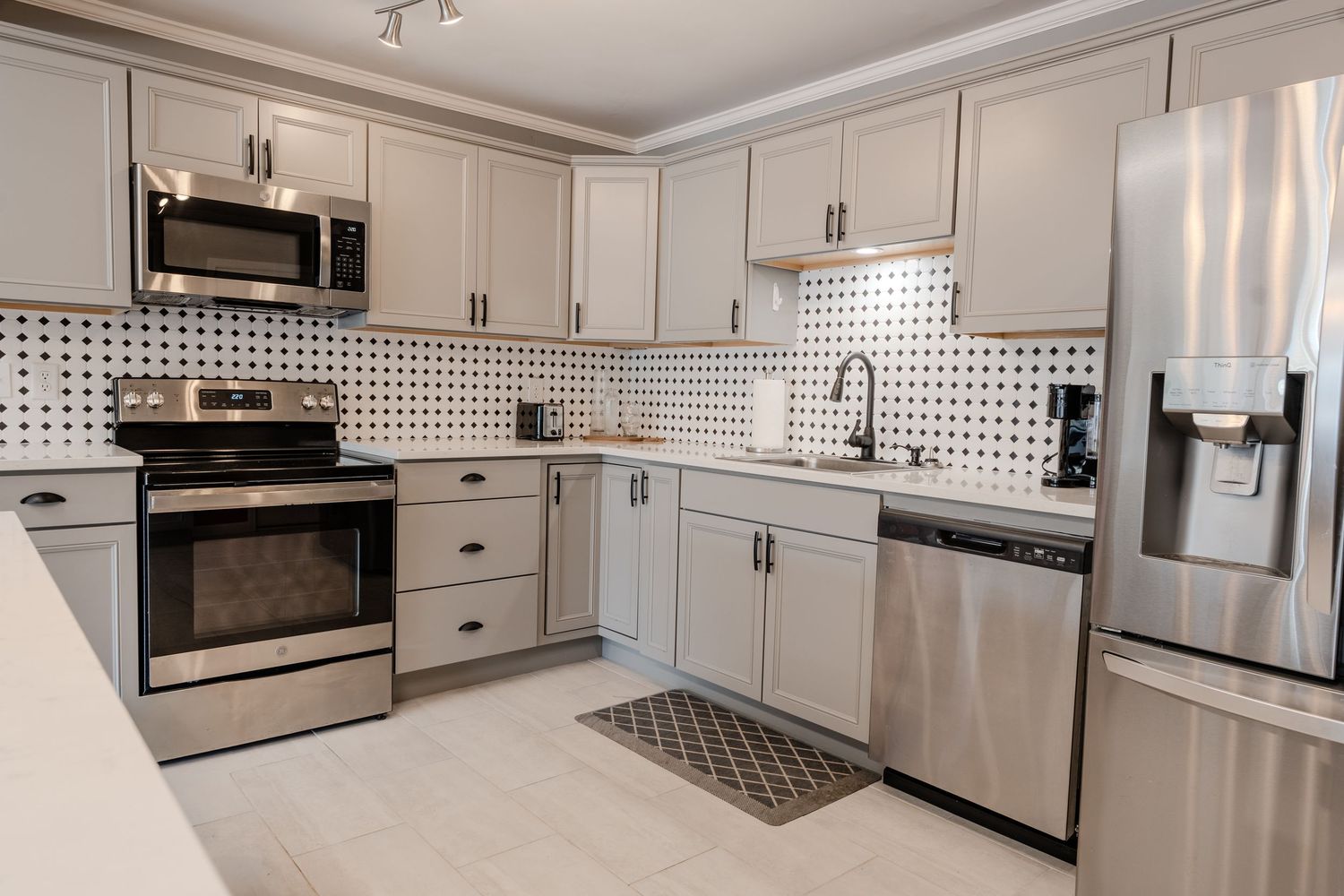 Modern white kitchen with stainless steel appliances, quartz countertops, and black and white geometric backsplash tile.