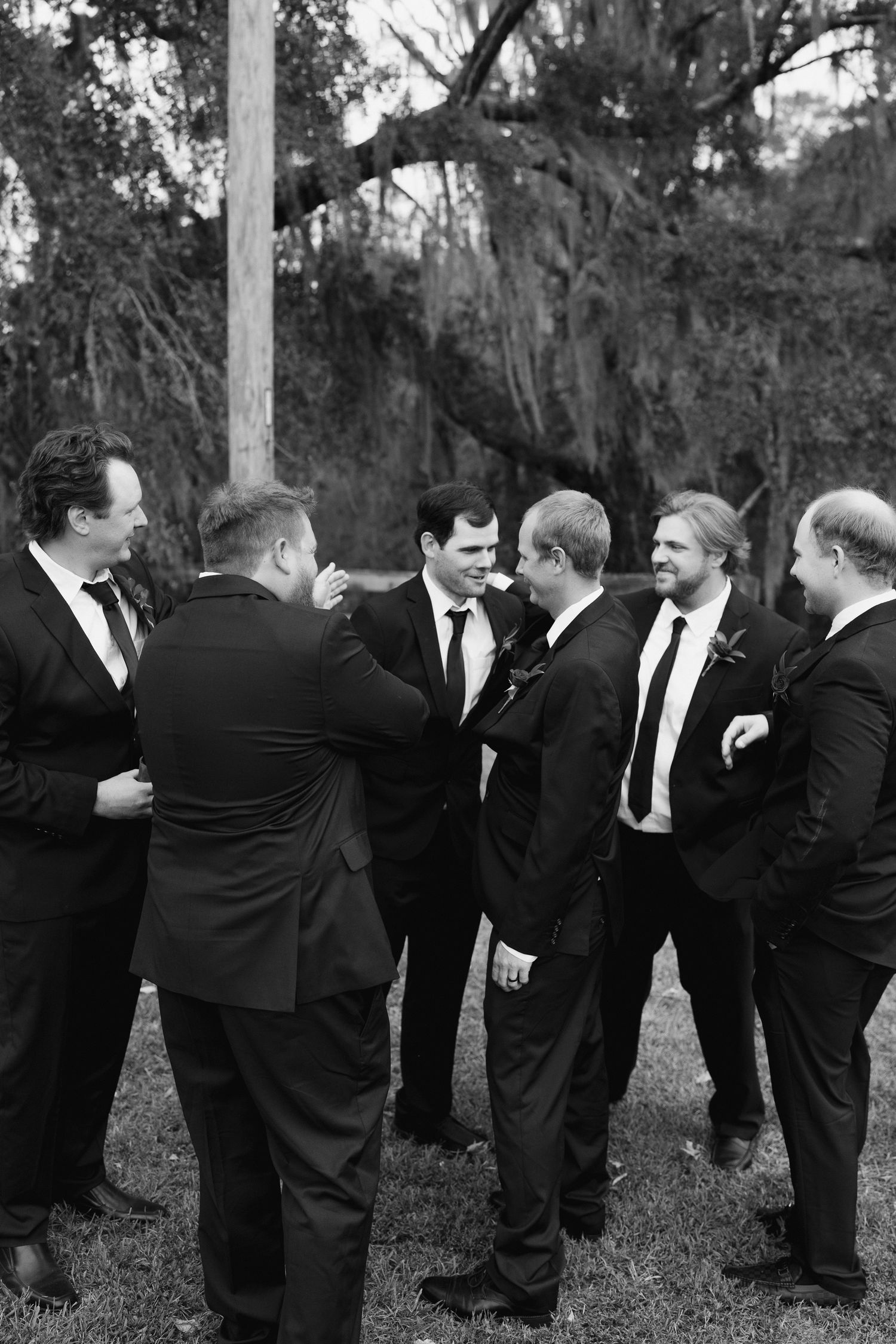 Groomsmen in black suits pose playfully in a line outdoors under moss-covered trees at a wedding.