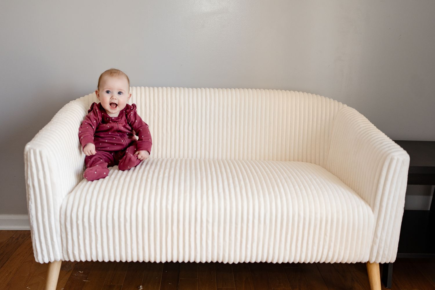 Baby in burgundy clothing exploring a white ribbed couch in a minimalist setting.