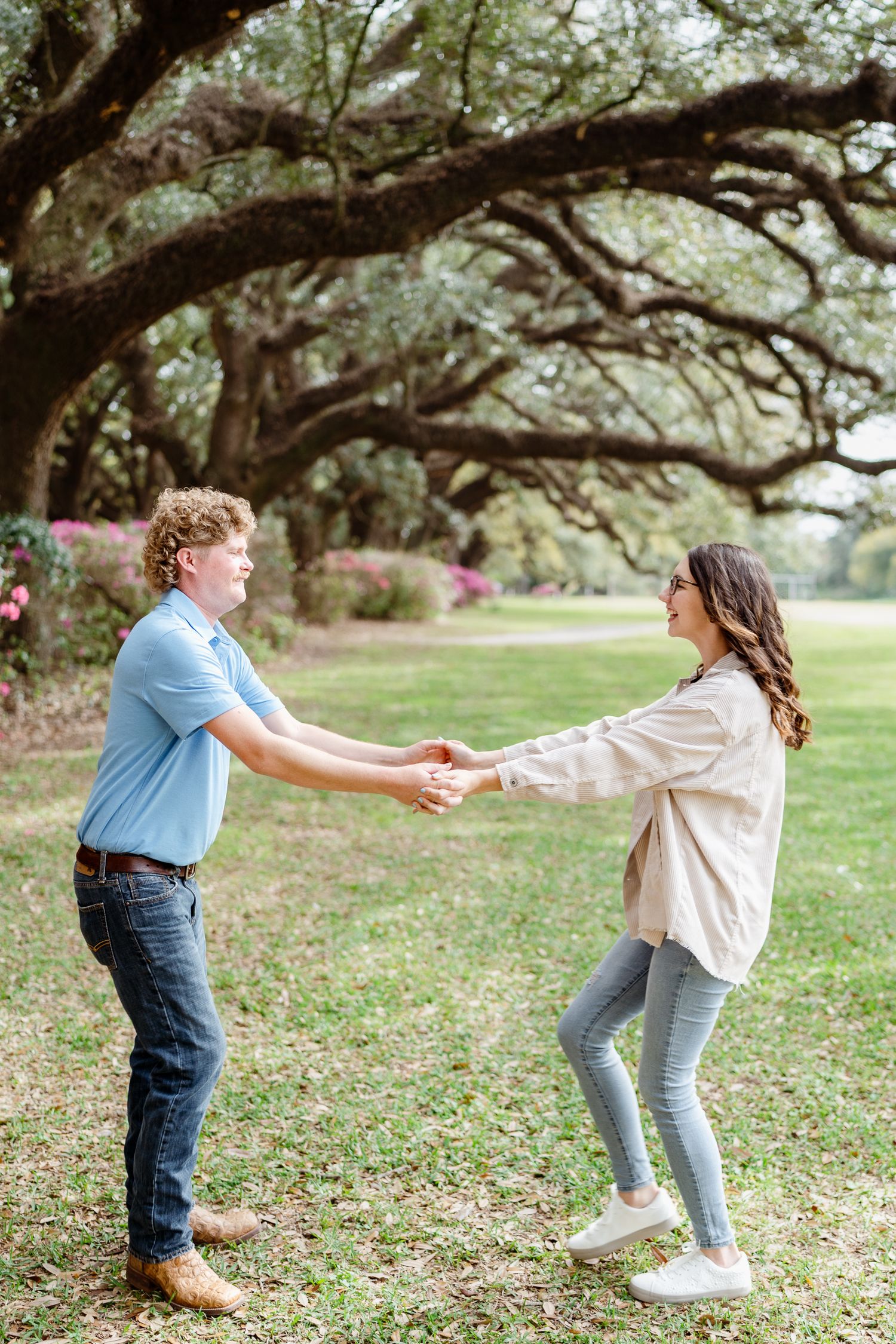 Two people dancing playfully on grass beneath oak trees.