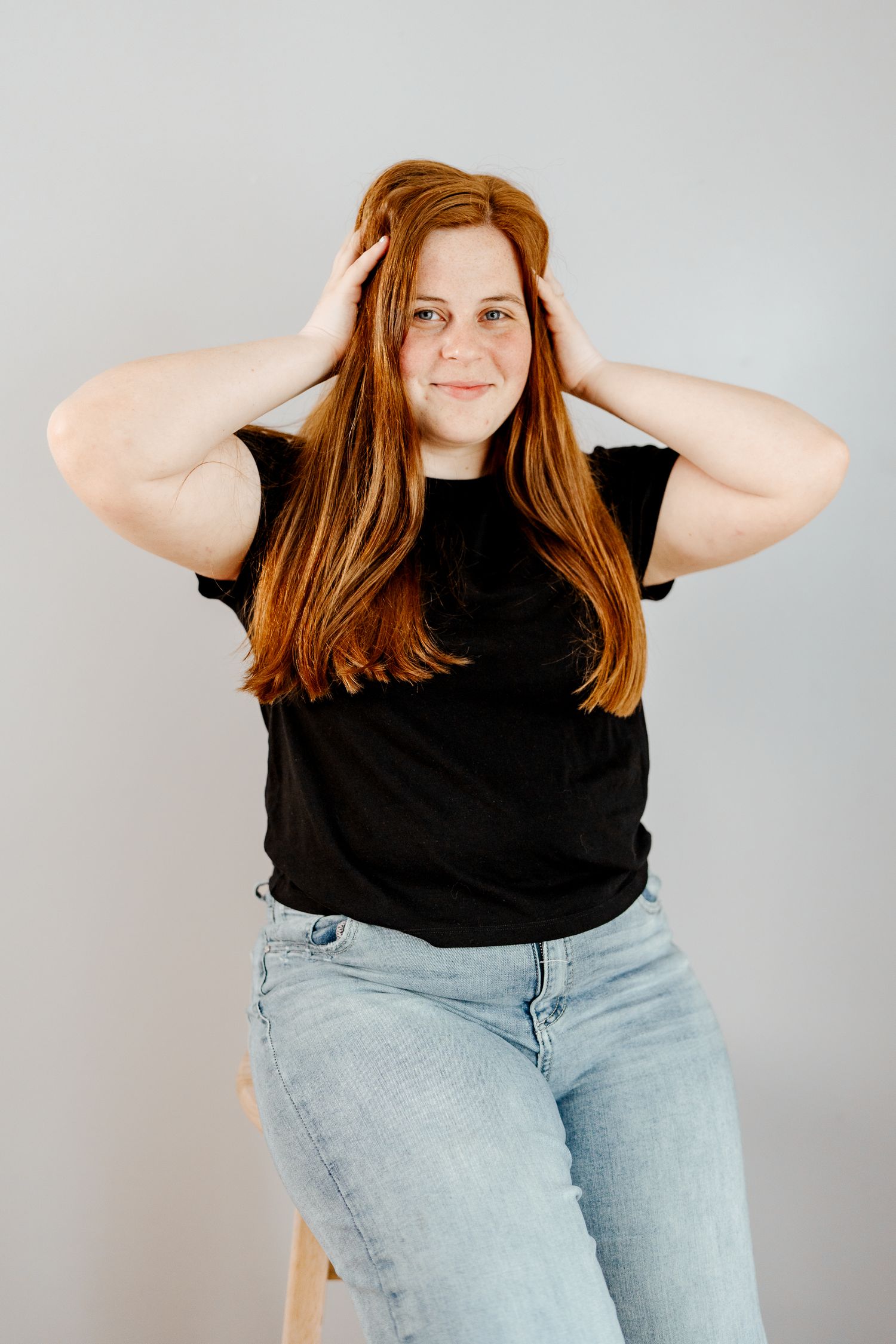 Person wearing black t-shirt and light denim poses playfully with hands in hair against grey backdrop.