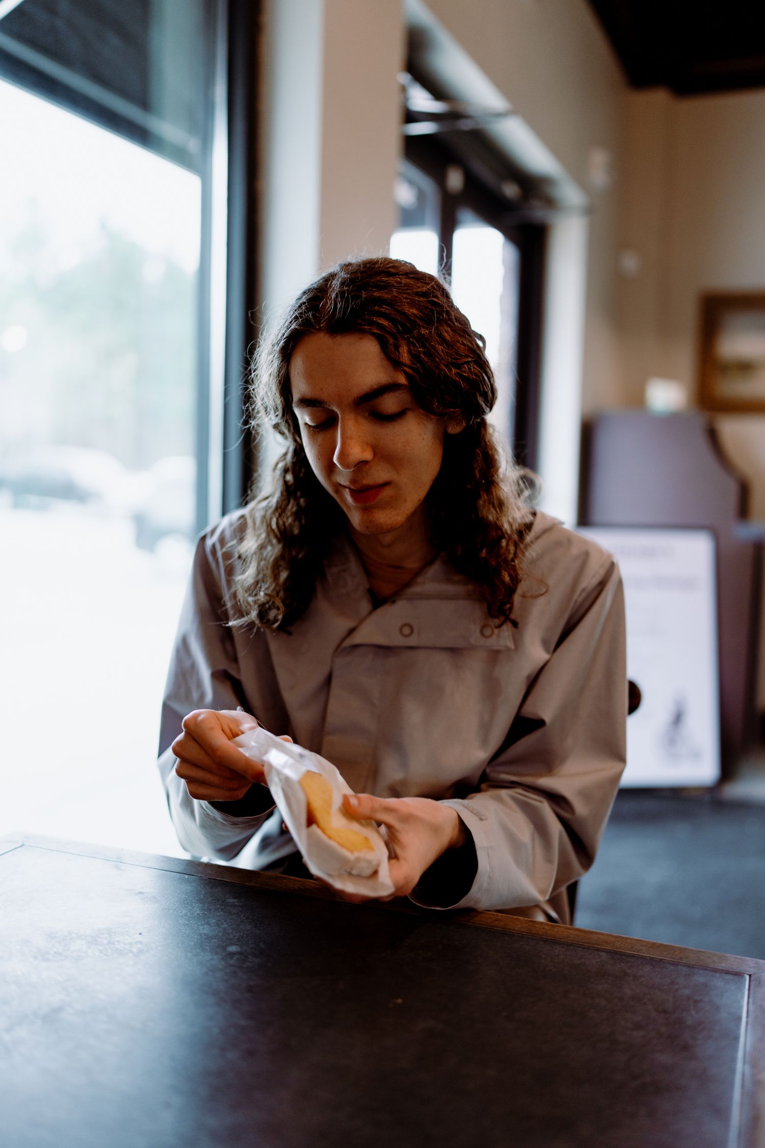 Person unwrapping food item while seated at dark wooden table by window.
