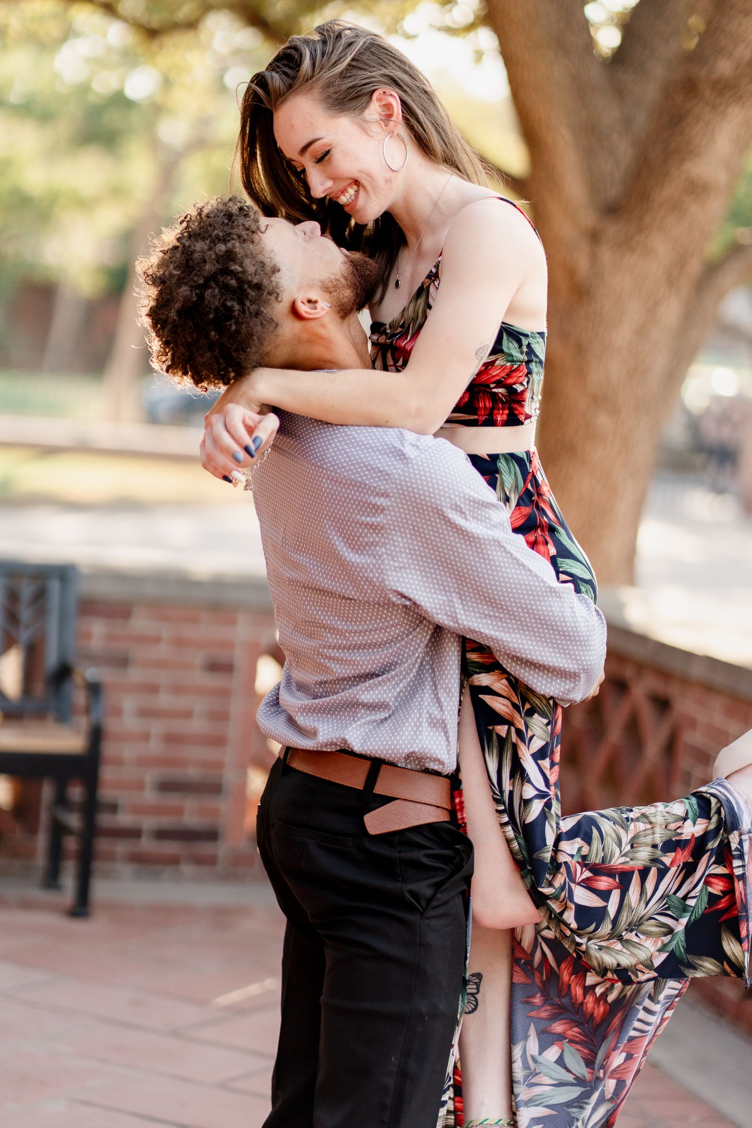 A romantic couple embraces and dances together during a sunset photoshoot in front of brick walls and trees.