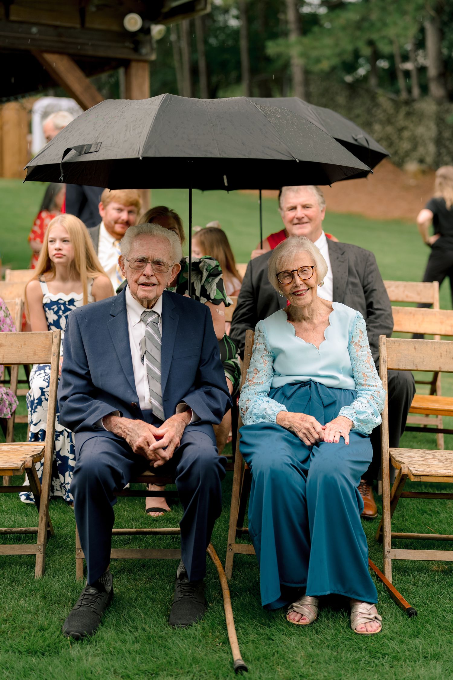Elderly couple seated at outdoor wedding ceremony under black umbrella on wooden folding chairs.