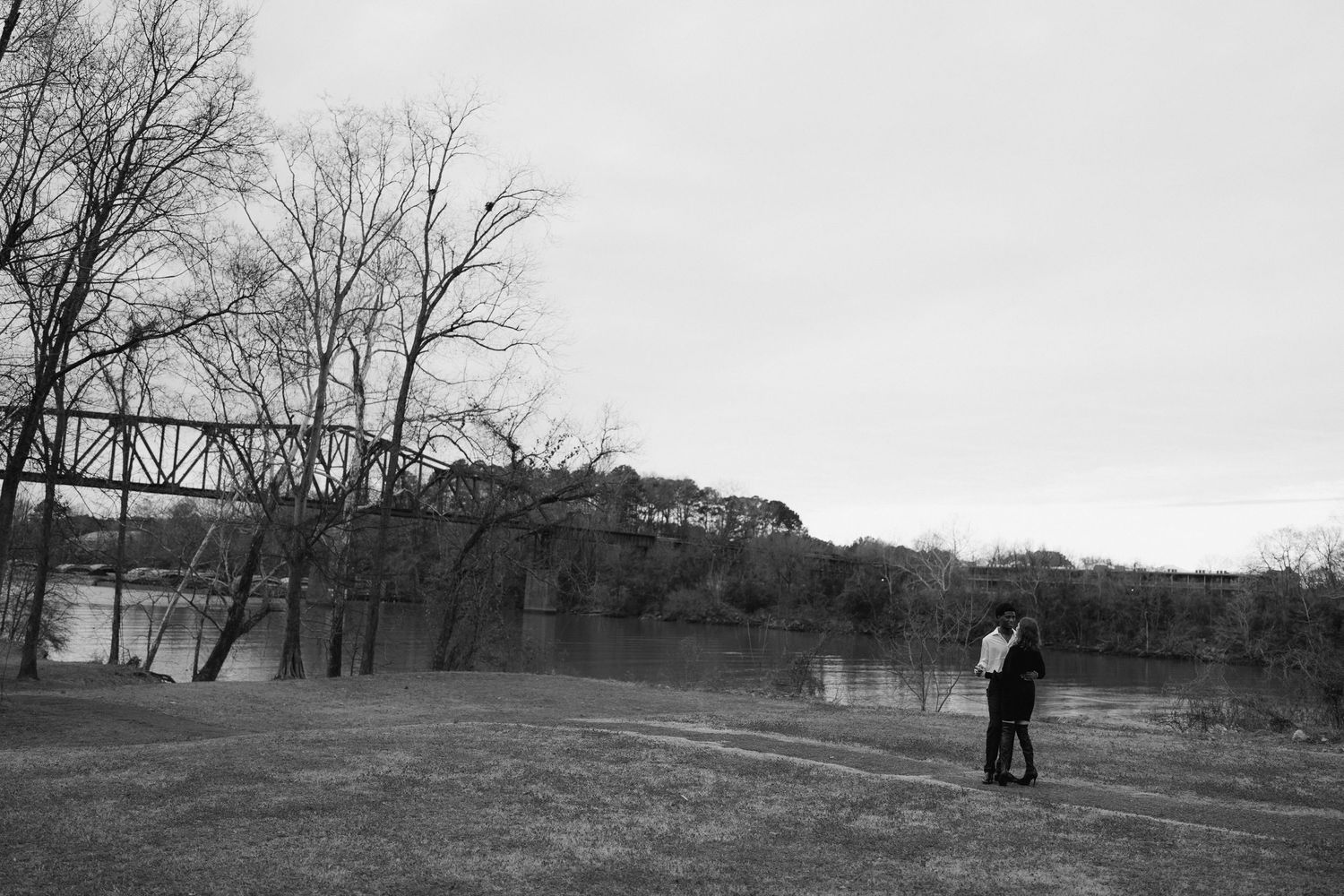 Black and white photo of couple kissing near river with iron trestle bridge in background.