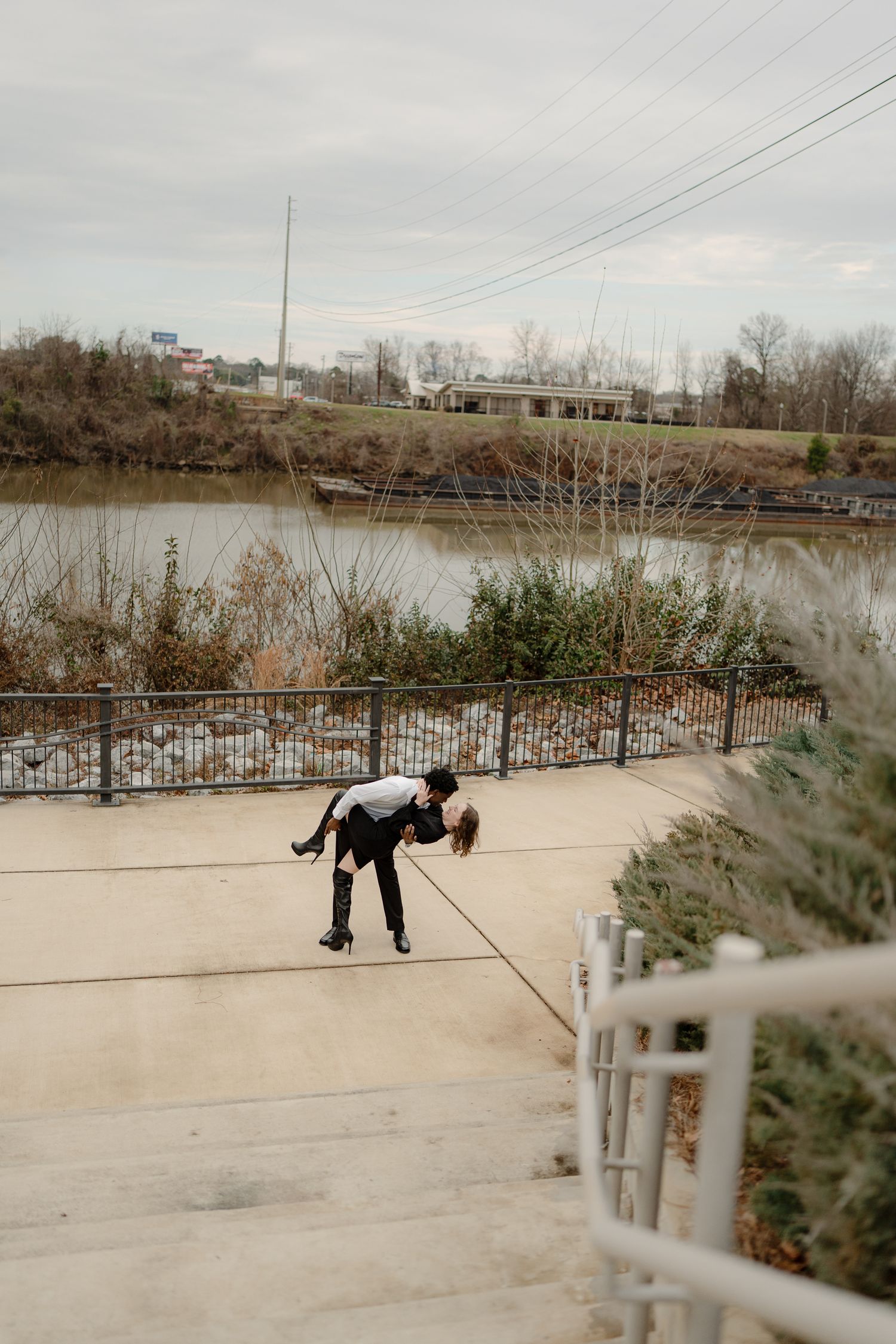 A couple shares a romantic dip pose on a waterfront promenade with a river and bare trees in the background.