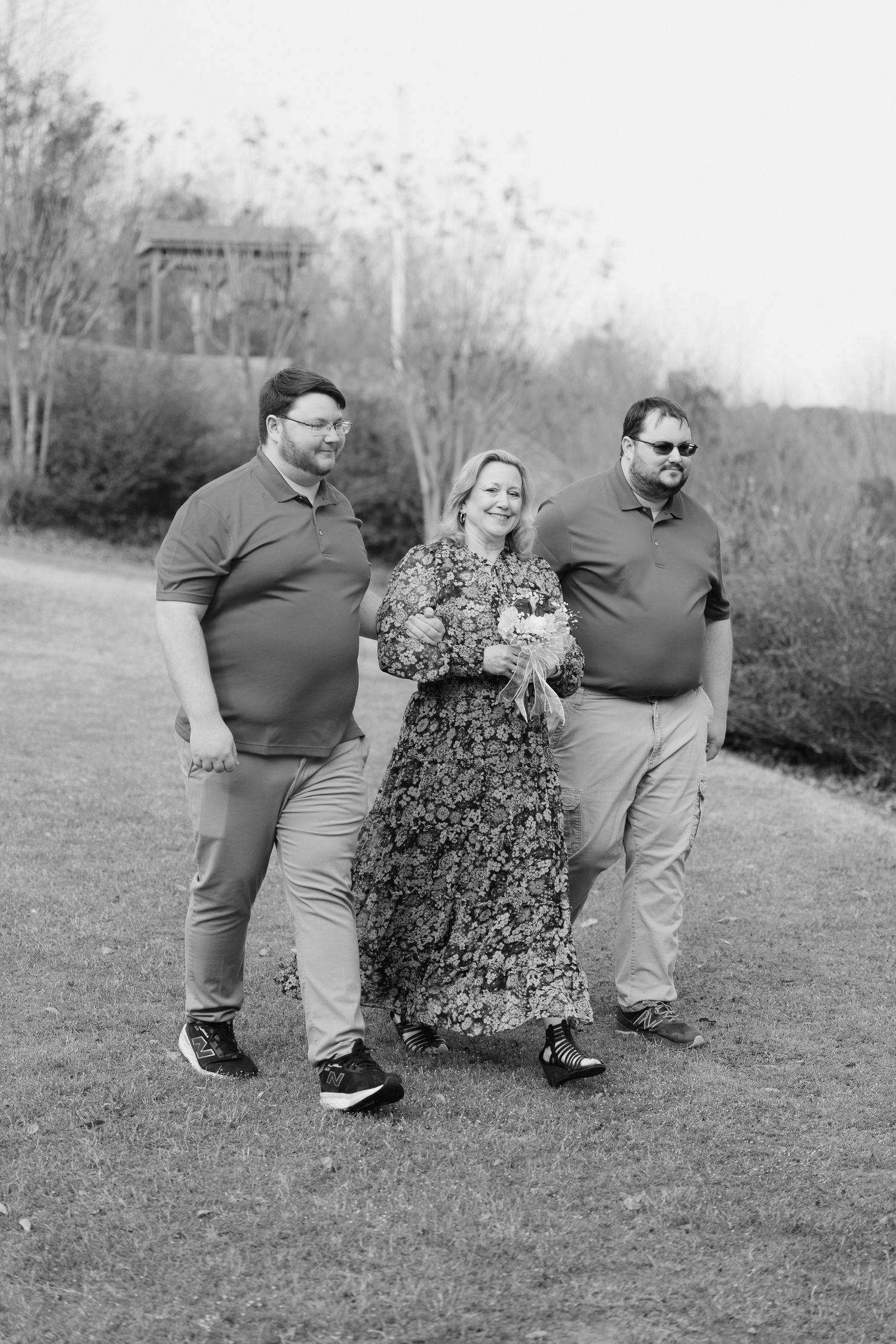 Black and white photo of a woman in floral dress walking with two men in polo shirts on a grassy path.