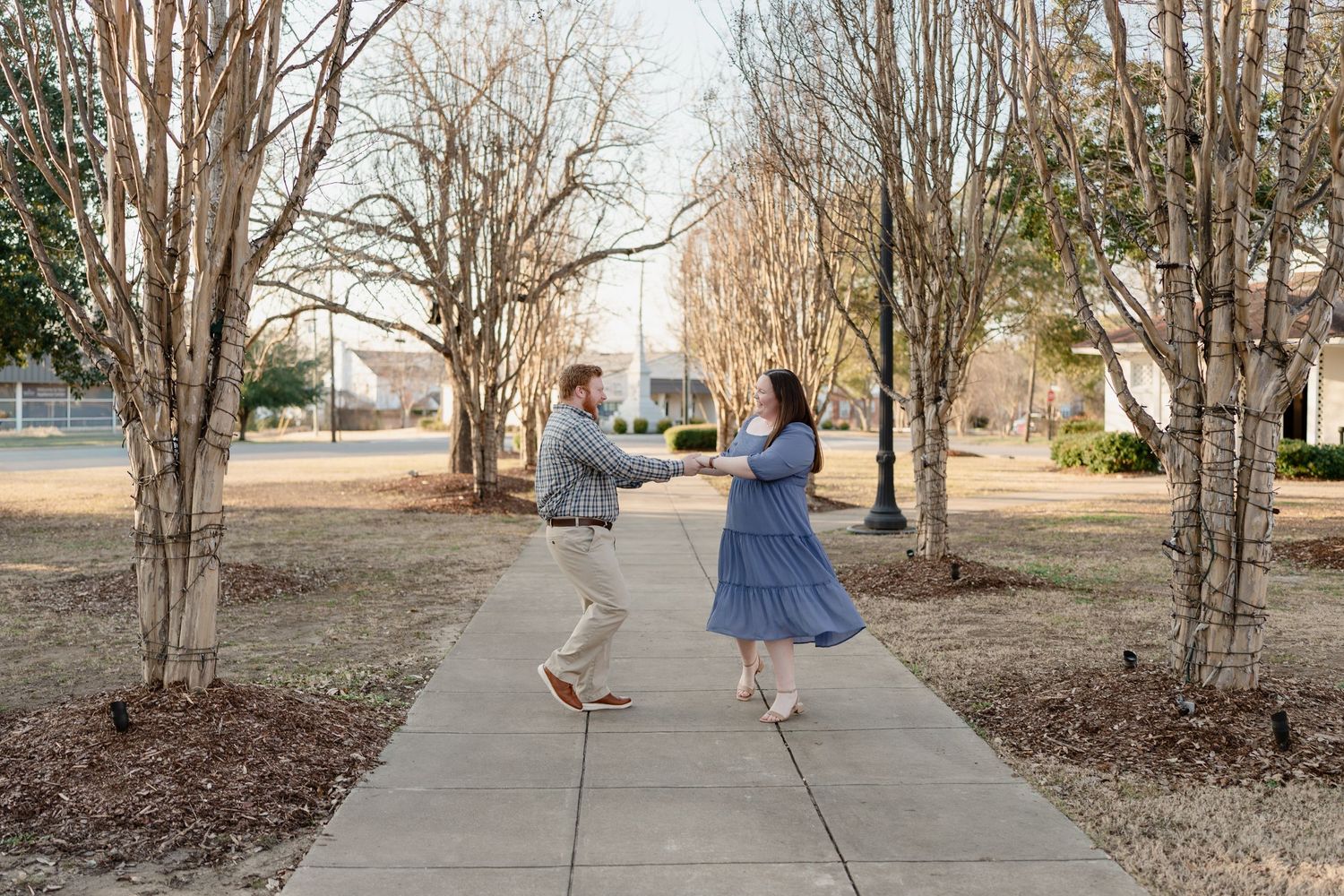 Couple holding hands on a sidewalk path surrounded by bare winter trees in a park setting.