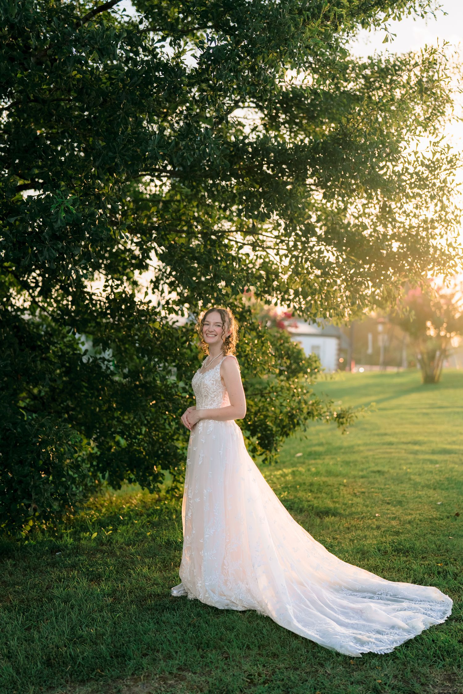 Bride in elegant lace wedding gown with long train stands under green tree in golden sunset light.
