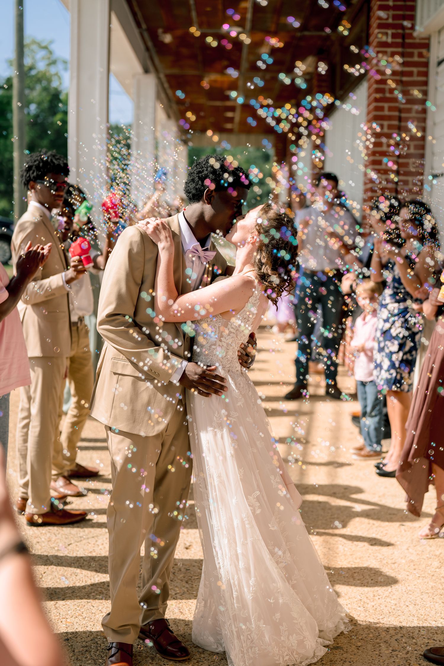 Newlywed couple shares kiss surrounded by colorful bubbles as guests celebrate their outdoor wedding send-off.