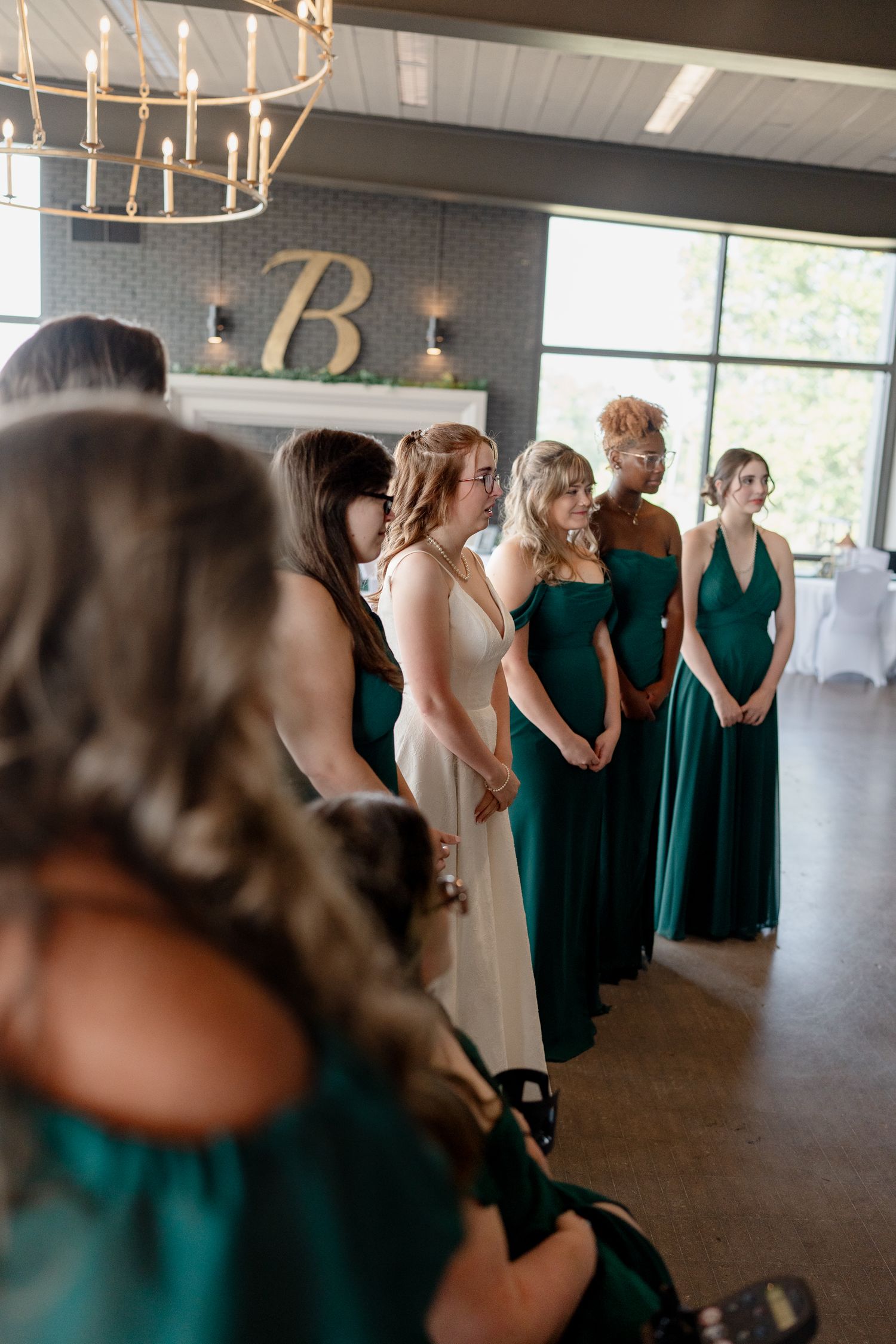 Wedding party in emerald green dresses gathered in an elegant venue with chandeliers.