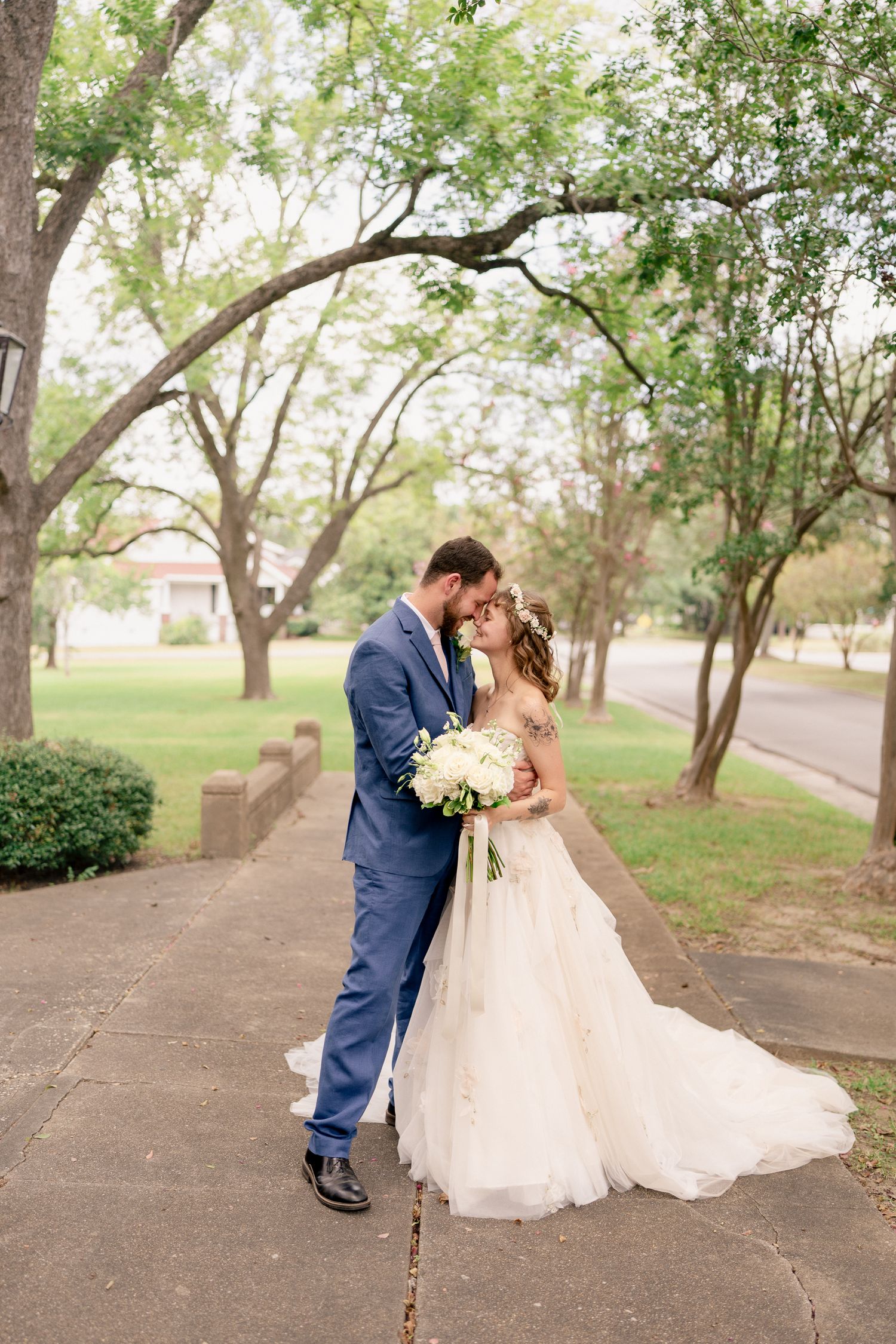 A romantic wedding couple shares an intimate moment on a tree-lined pathway in their elegant attire.