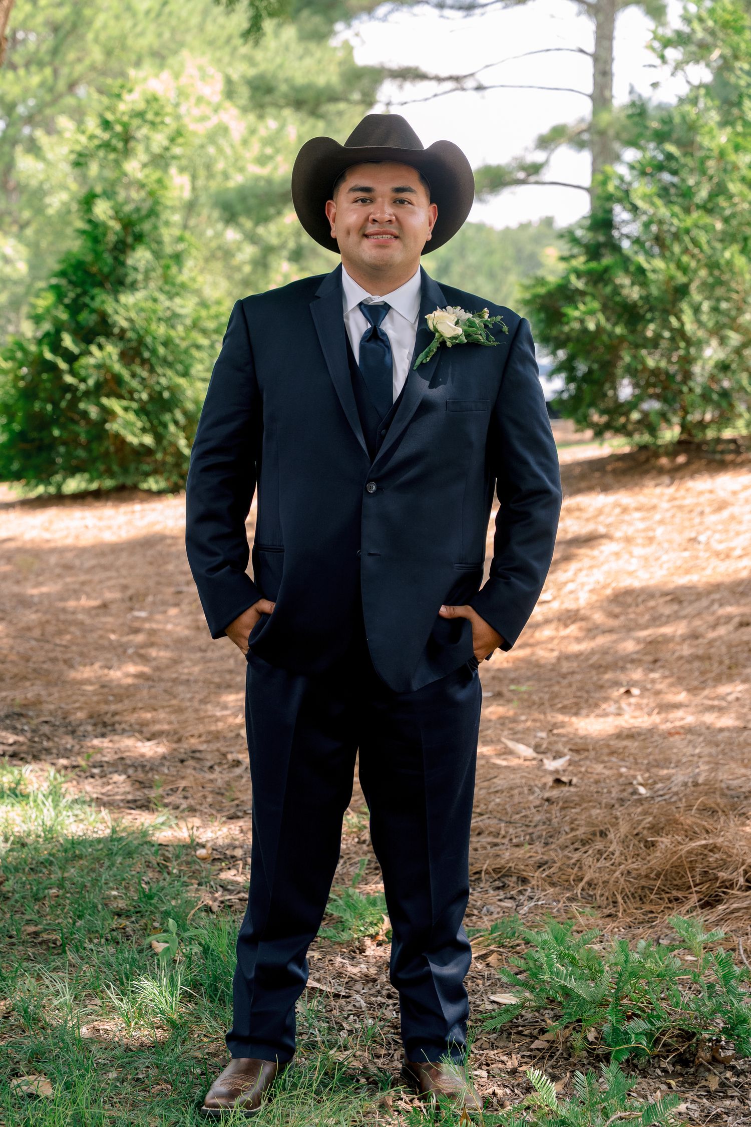 A person in a formal black suit and cowboy hat stands in an outdoor setting with trees and foliage in the background.