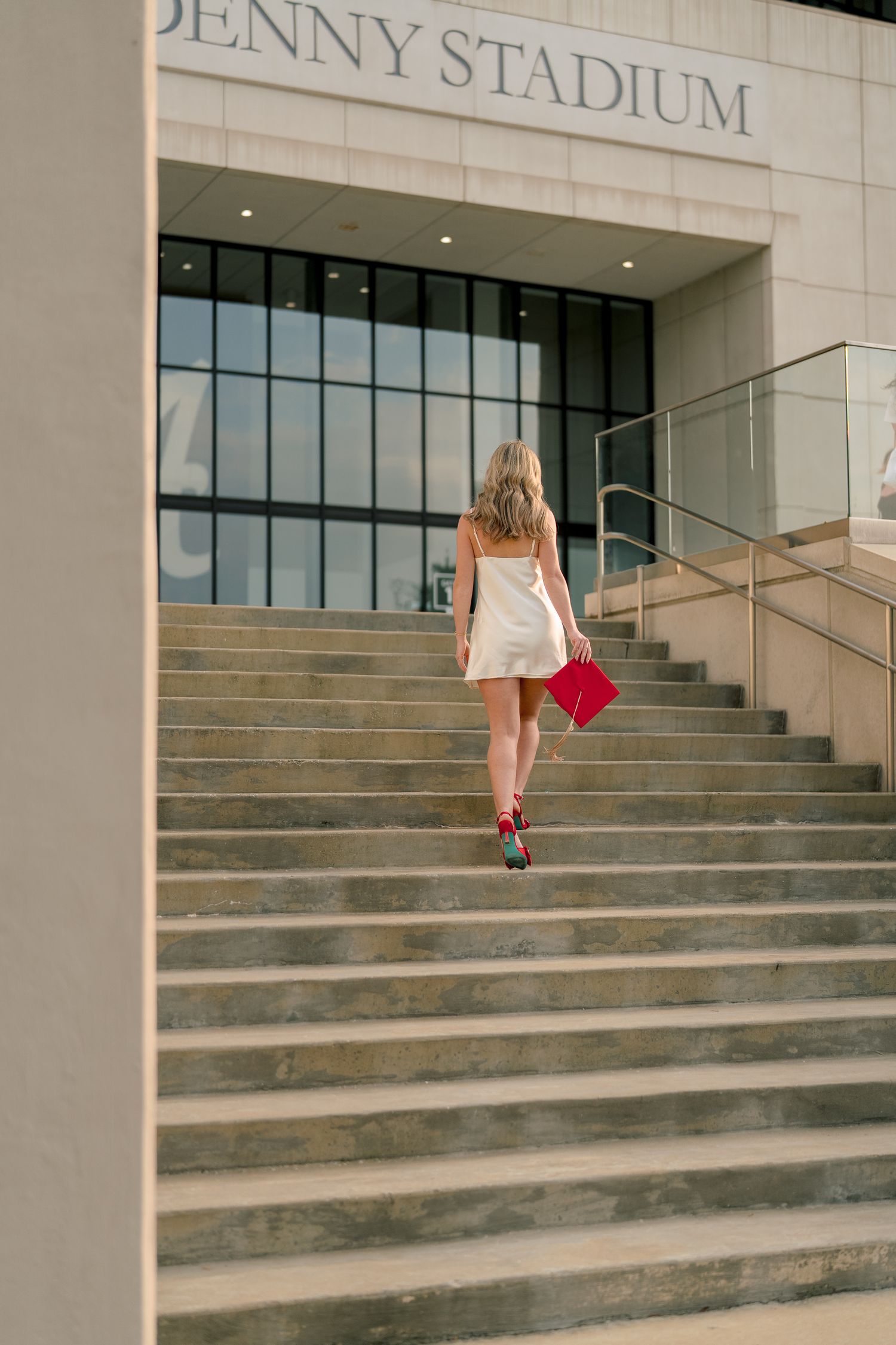 A person in white ascending the concrete steps of Denny Stadium while holding a red graduation cap.