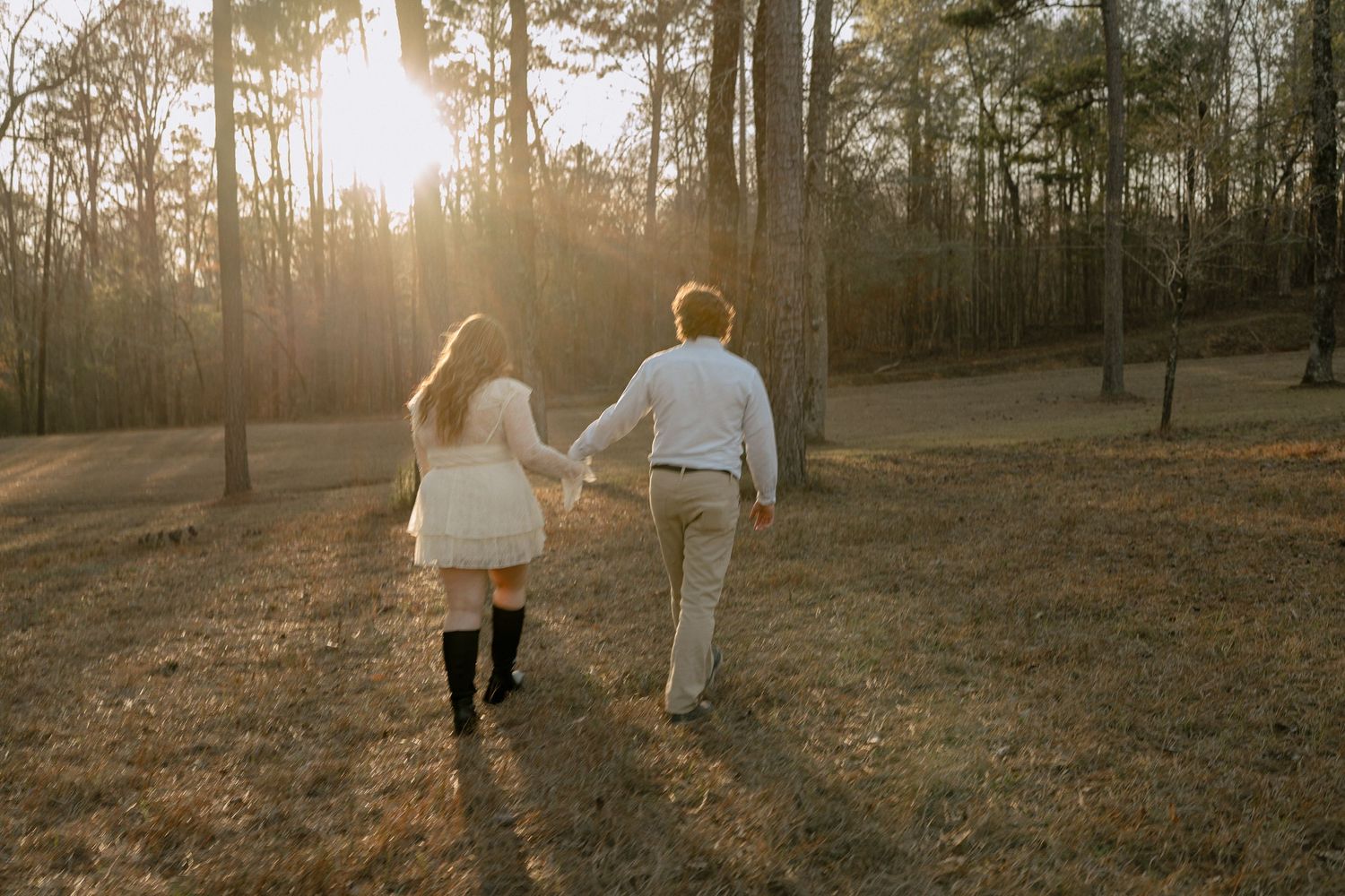 Couple walking hand in hand through a sun-drenched forest at golden hour, viewed from behind.