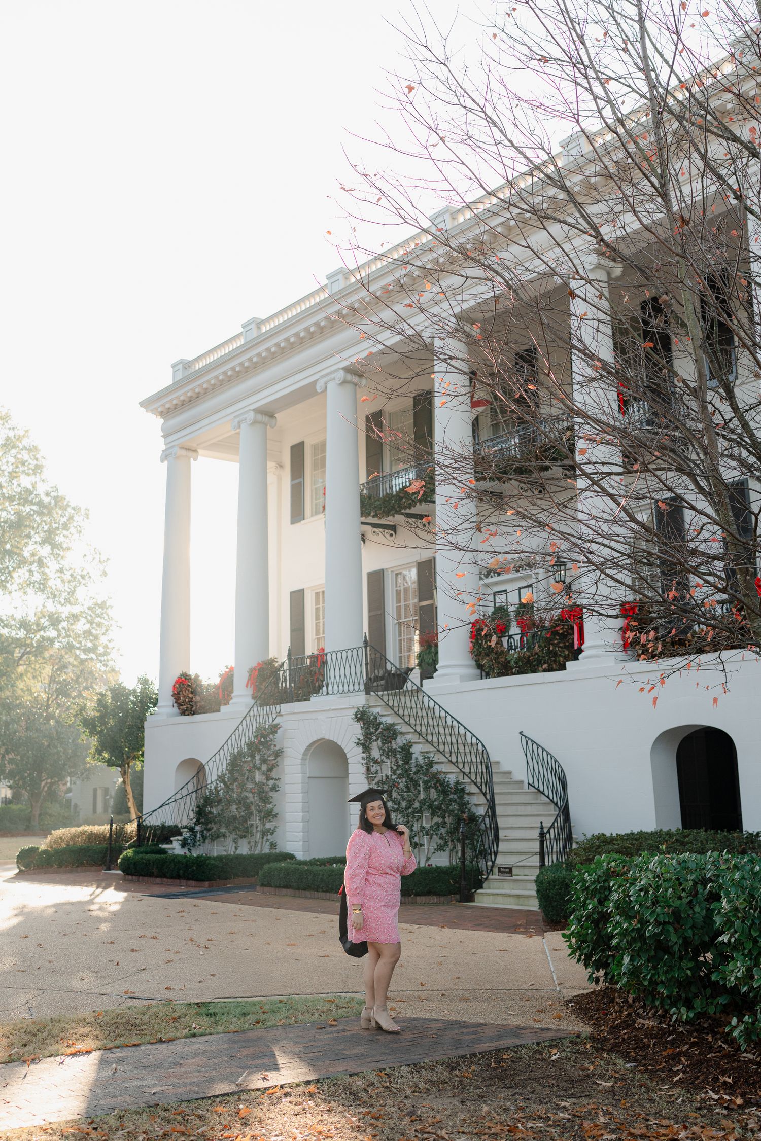 A graduate in pink dress poses in front of a grand white antebellum-style building with columns.
