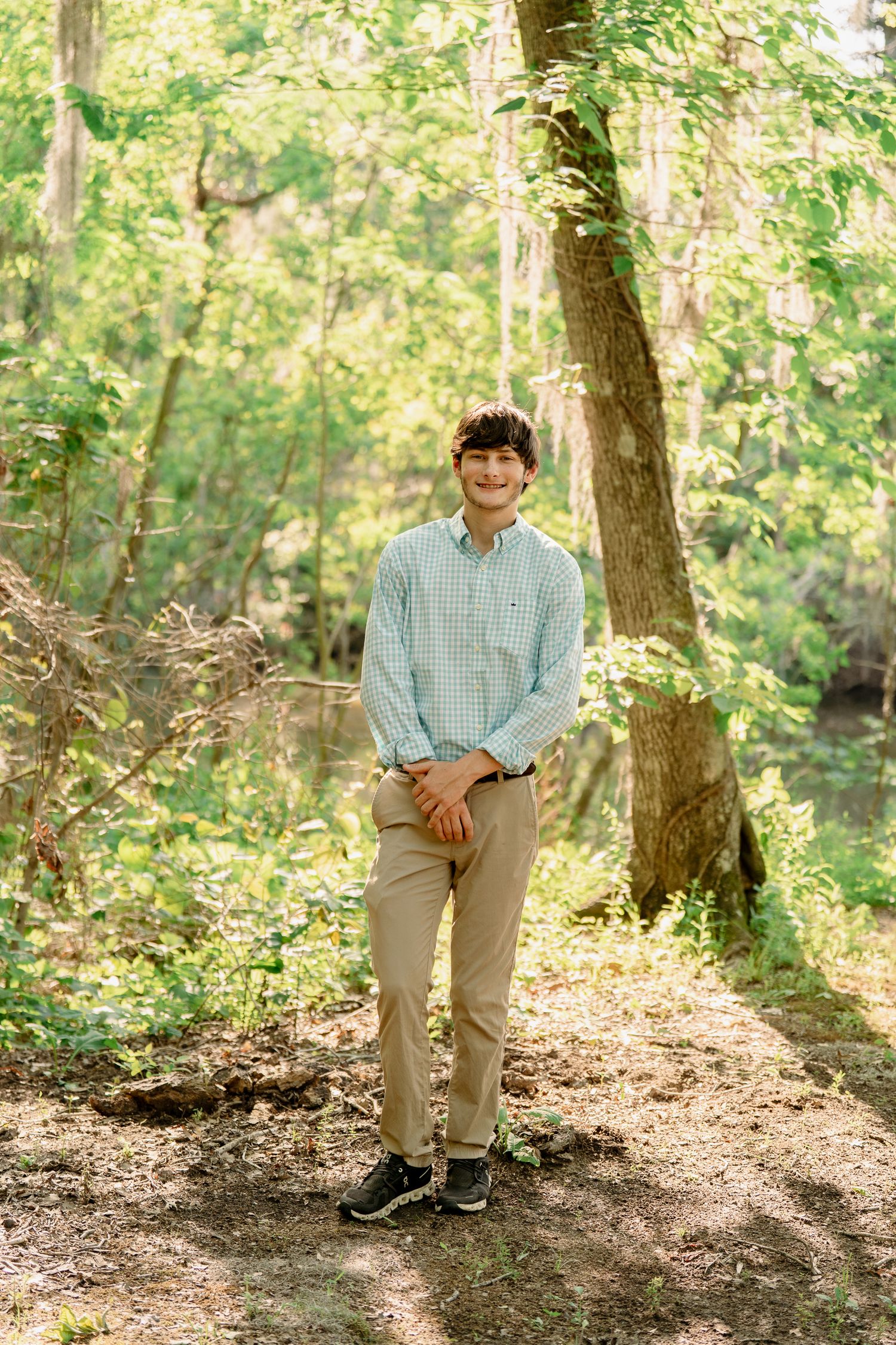 Student in light blue button-down shirt stands on forest path during photo session.