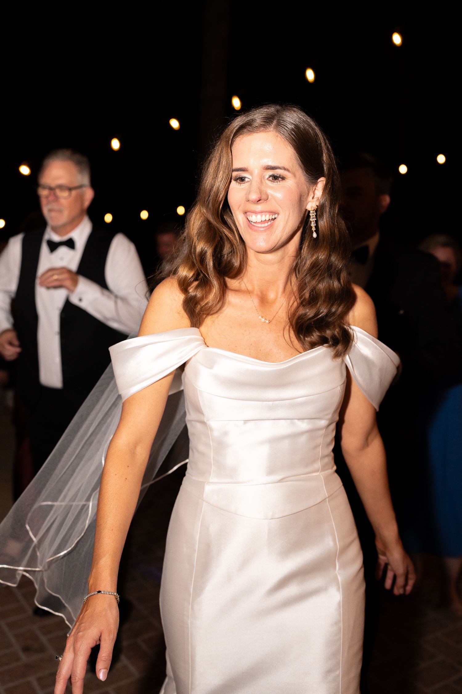 Woman in white satin dress dances joyfully under string lights at evening celebration.