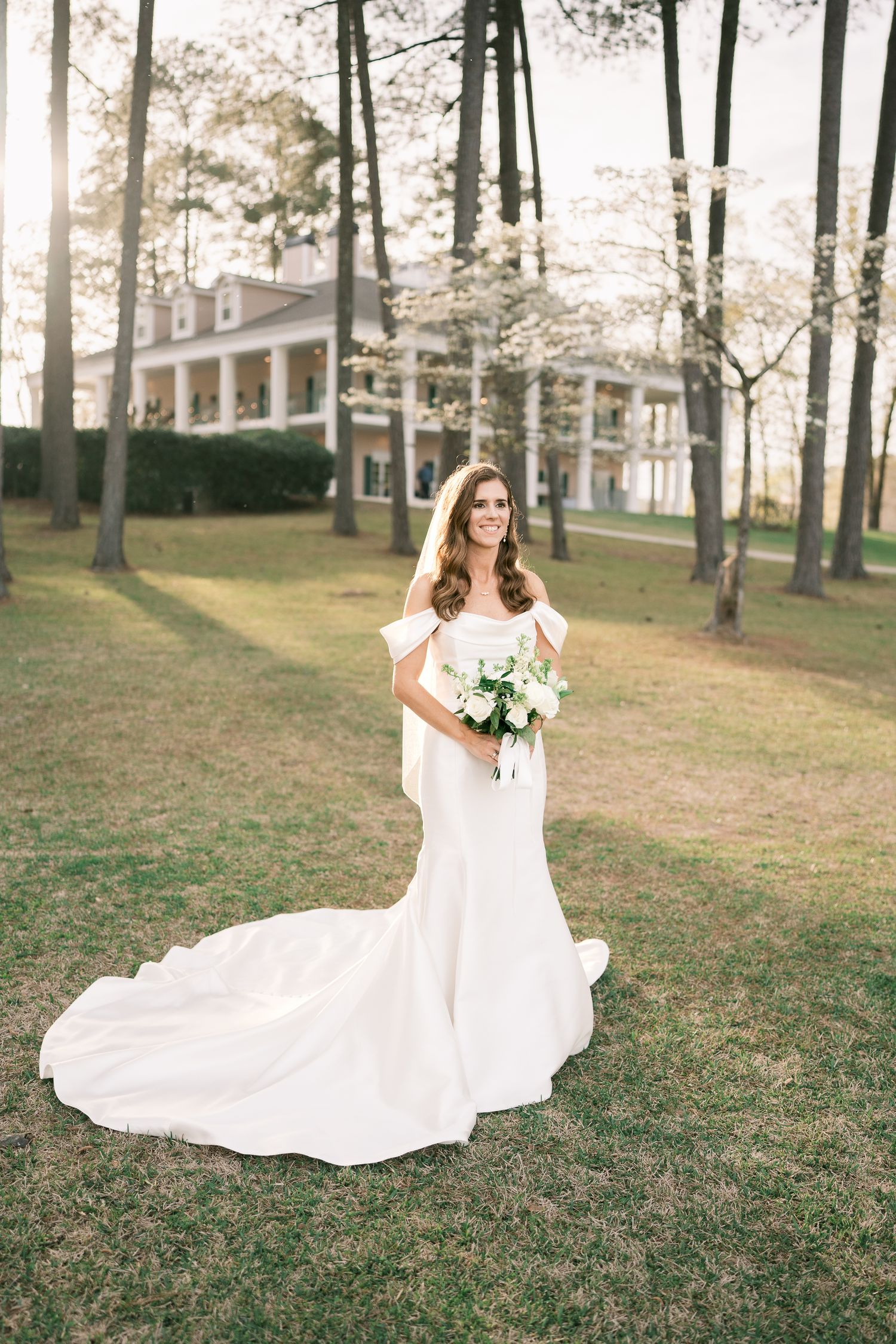 Bride in fitted white gown with train poses on lawn in front of Southern colonial mansion.