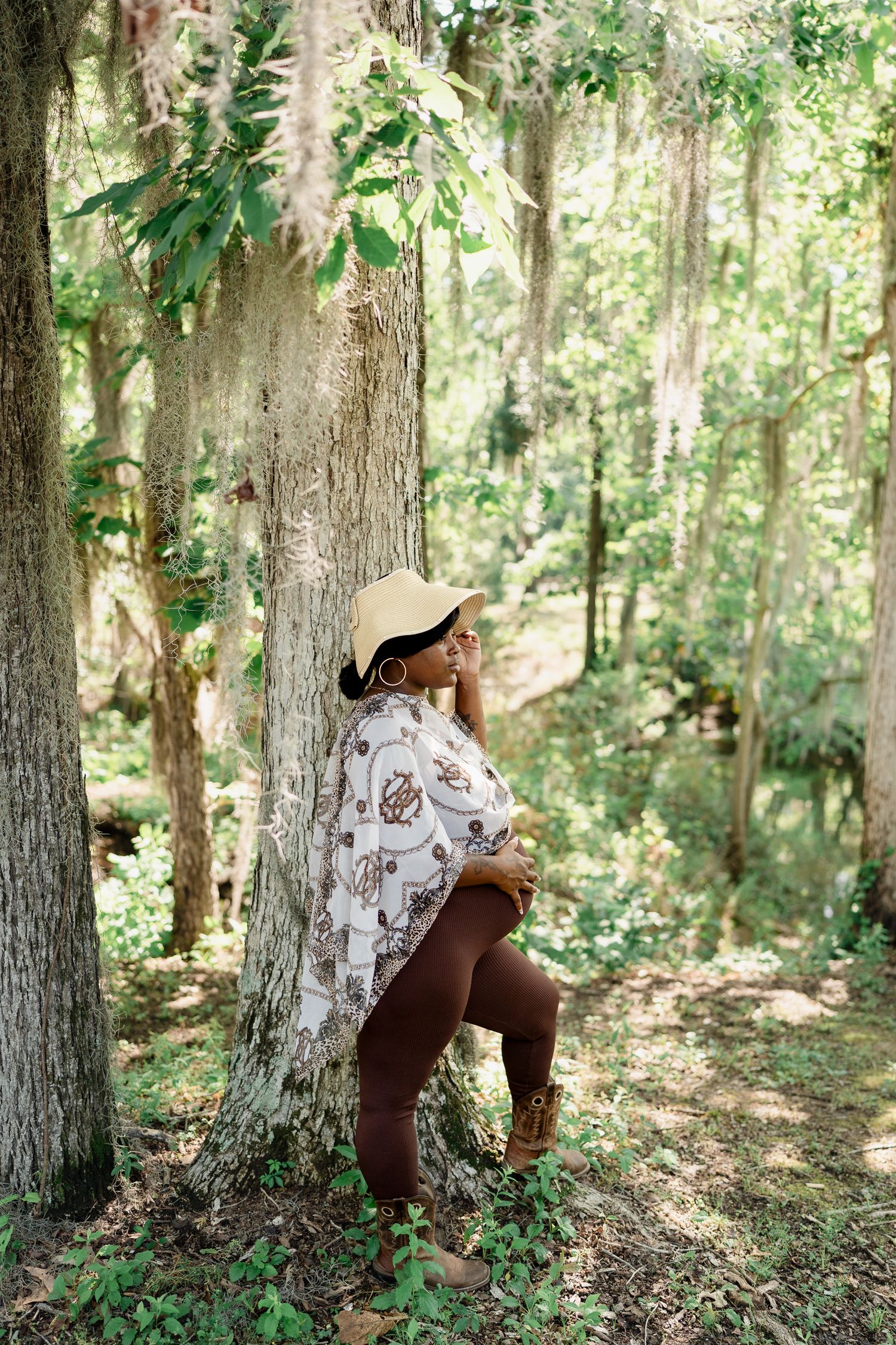 Person in flowing white poncho and sun hat poses dramatically against a tree in a serene forest setting.