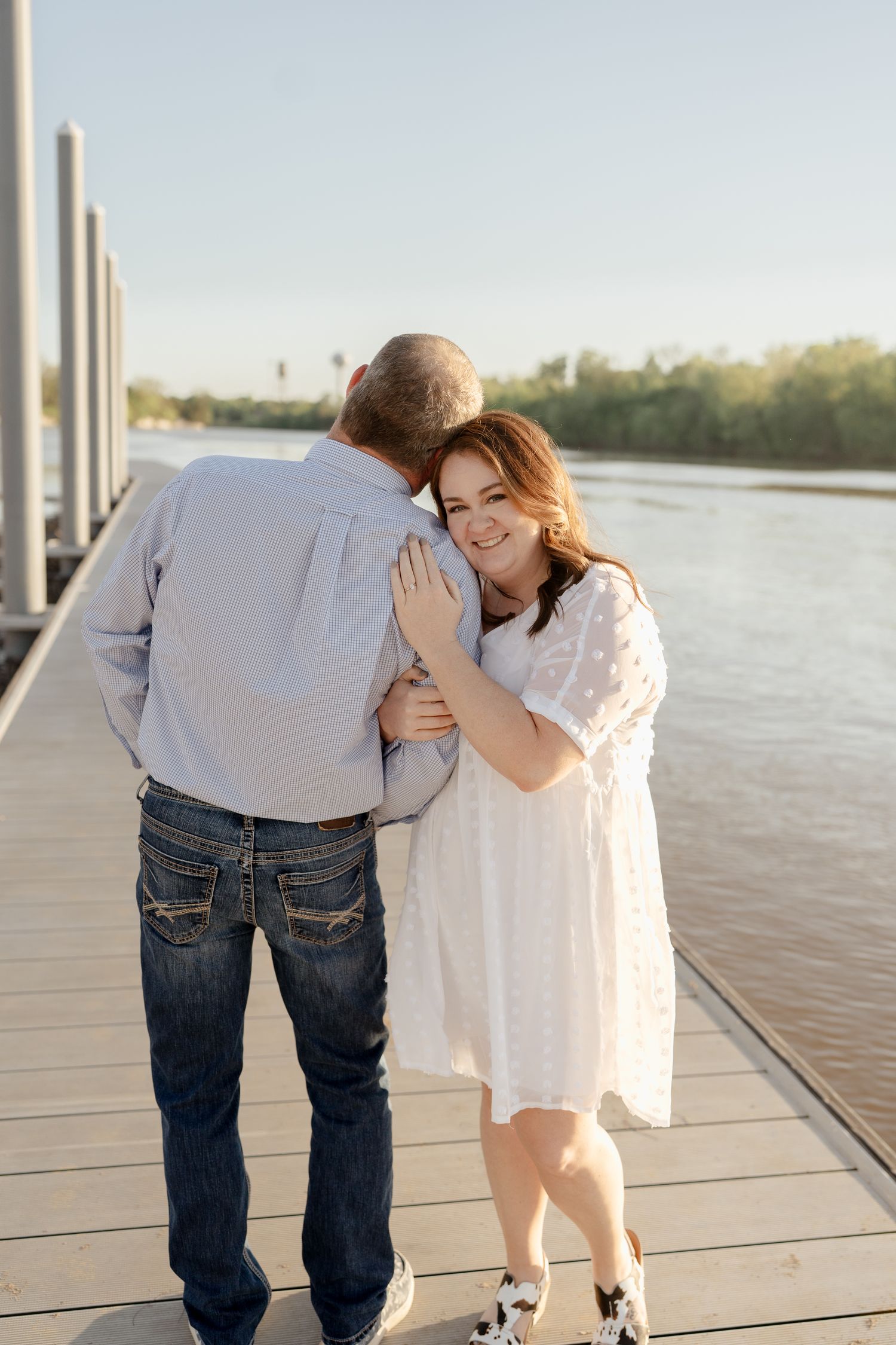 Two people share an affectionate moment while walking along a riverside dock at sunset.