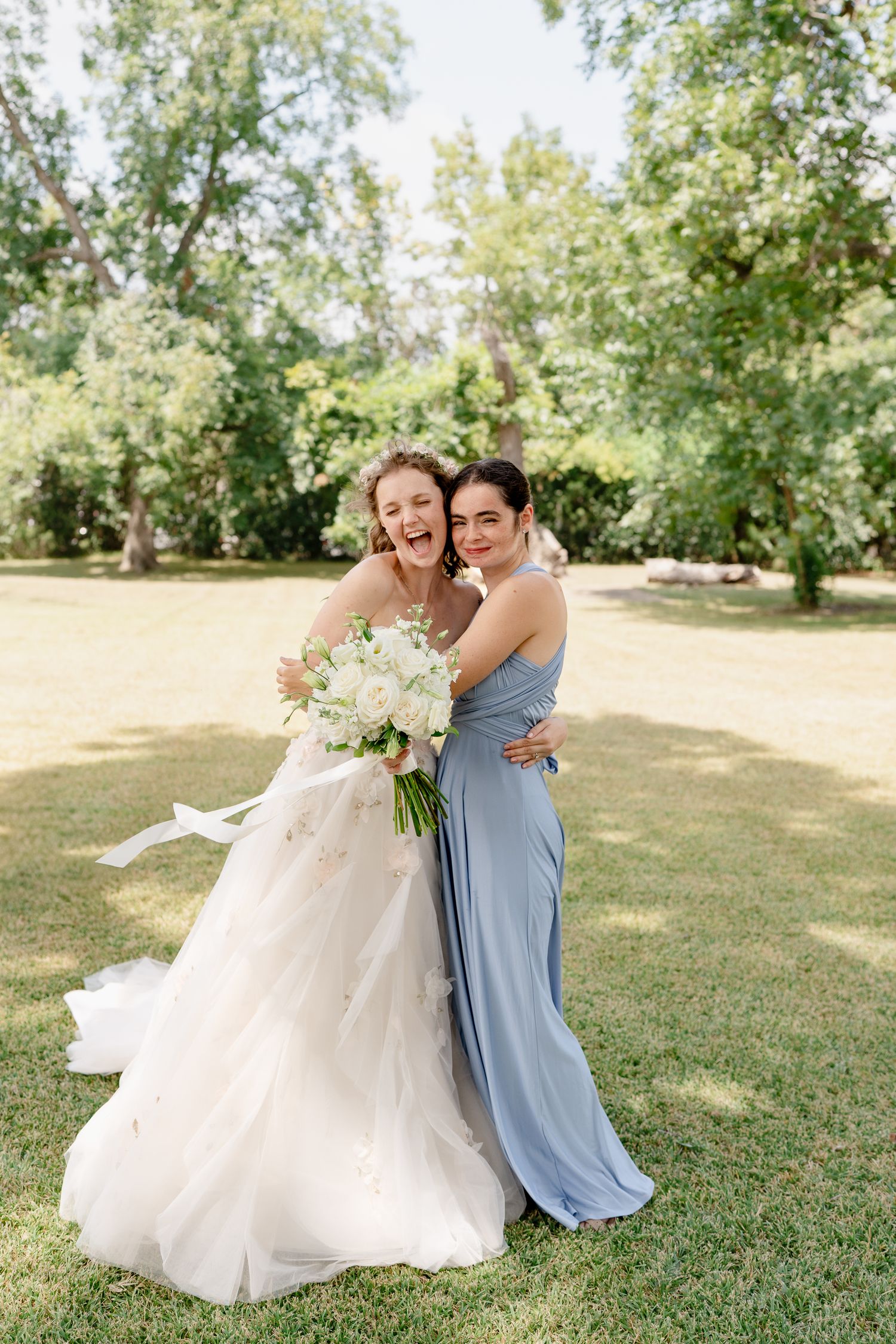 A bride in a flowing white gown and companion in blue embrace while holding white bouquets in a sunny garden setting.