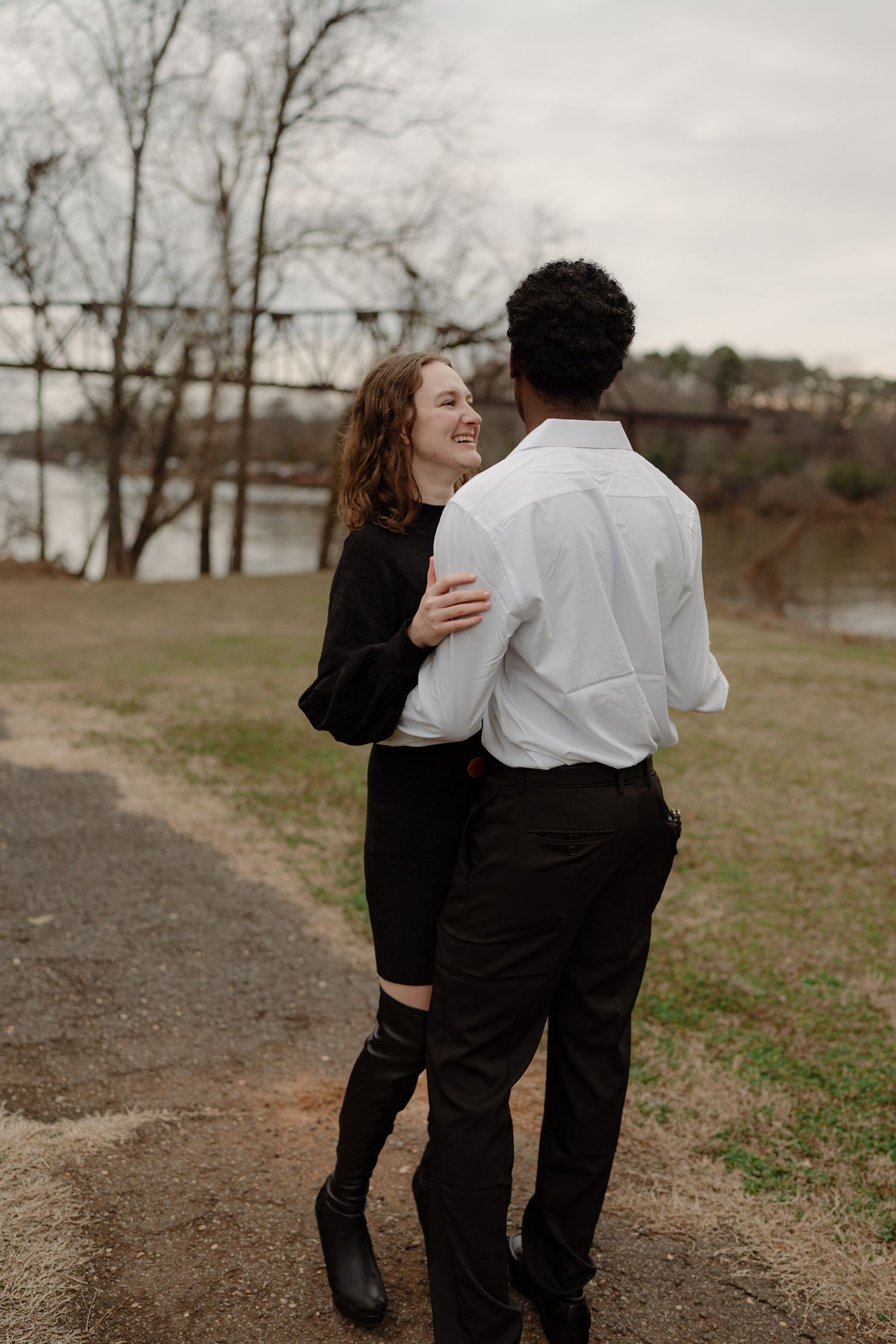 A smiling couple embraces outdoors, woman in black dress and man in white shirt, with a bridge and river in the background.