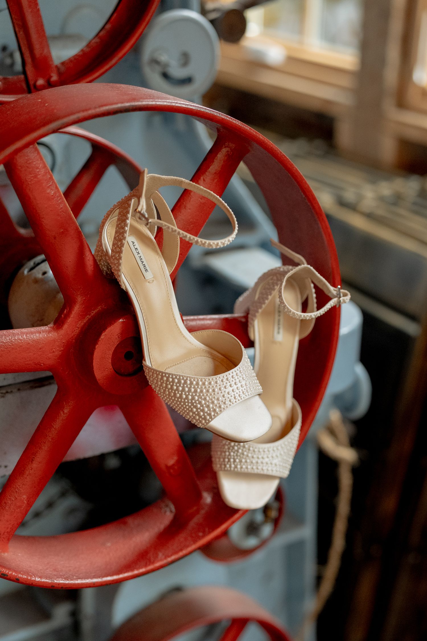 Beige pearl-embellished bridal heels with ankle straps displayed on a vintage red industrial wheel.